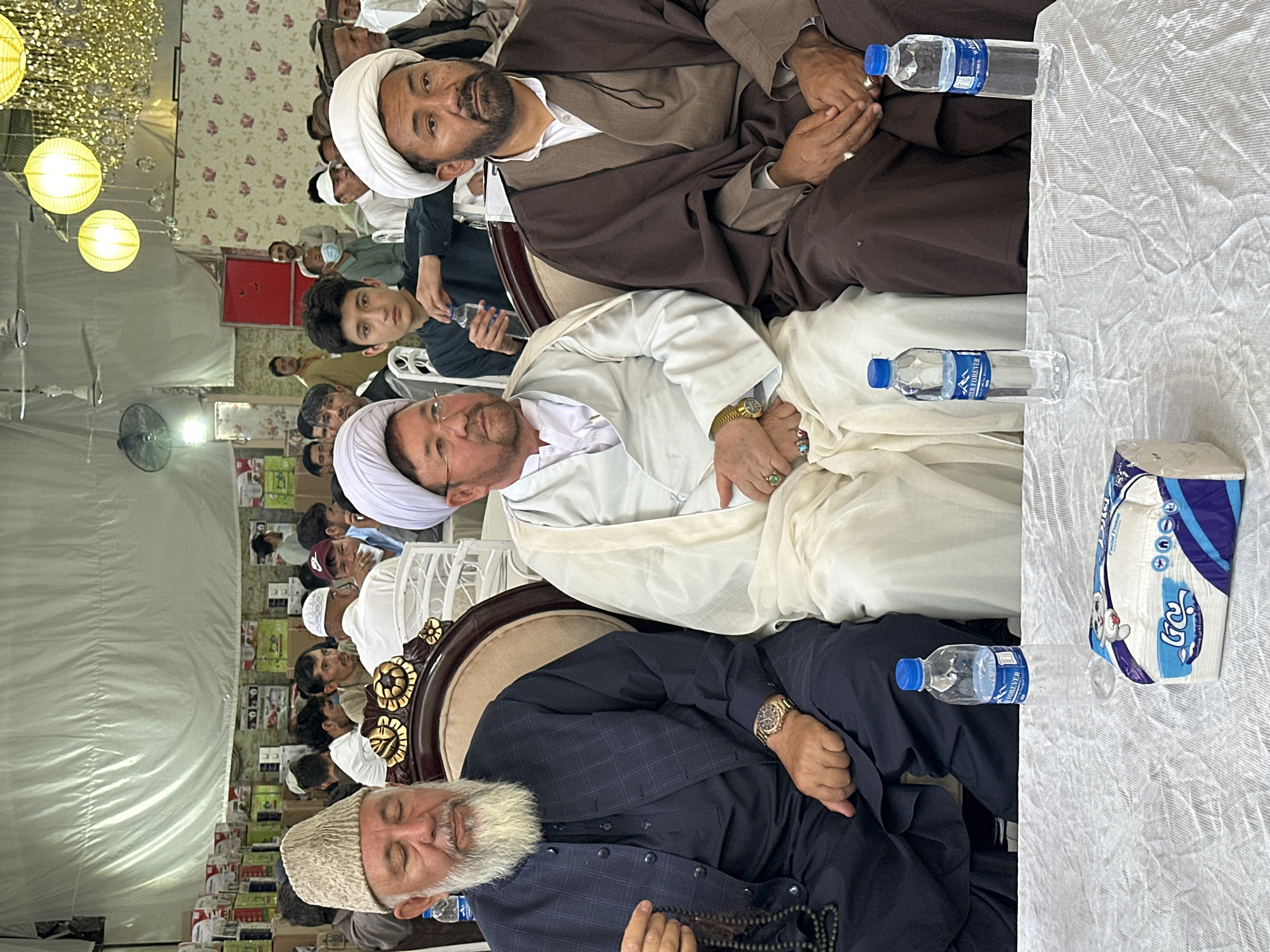 The image shows three men in religious attire sitting in chairs at a formal event, possibly in Quetta City Tehsil, Pakistan. The men are wearing traditional clothing, including white and brown robes and turbans. A younger boy is sitting behind the men, holding a water bottle. A table with bottled water and a tissue box is in the right foreground. Other individuals are visible in the background, seemingly attending the same gathering. The environment appears to be indoors, possibly a tent or hall, with floral wallpaper, chandeliers, and fans. There are boxes or packages stacked along a wall behind the attendees.