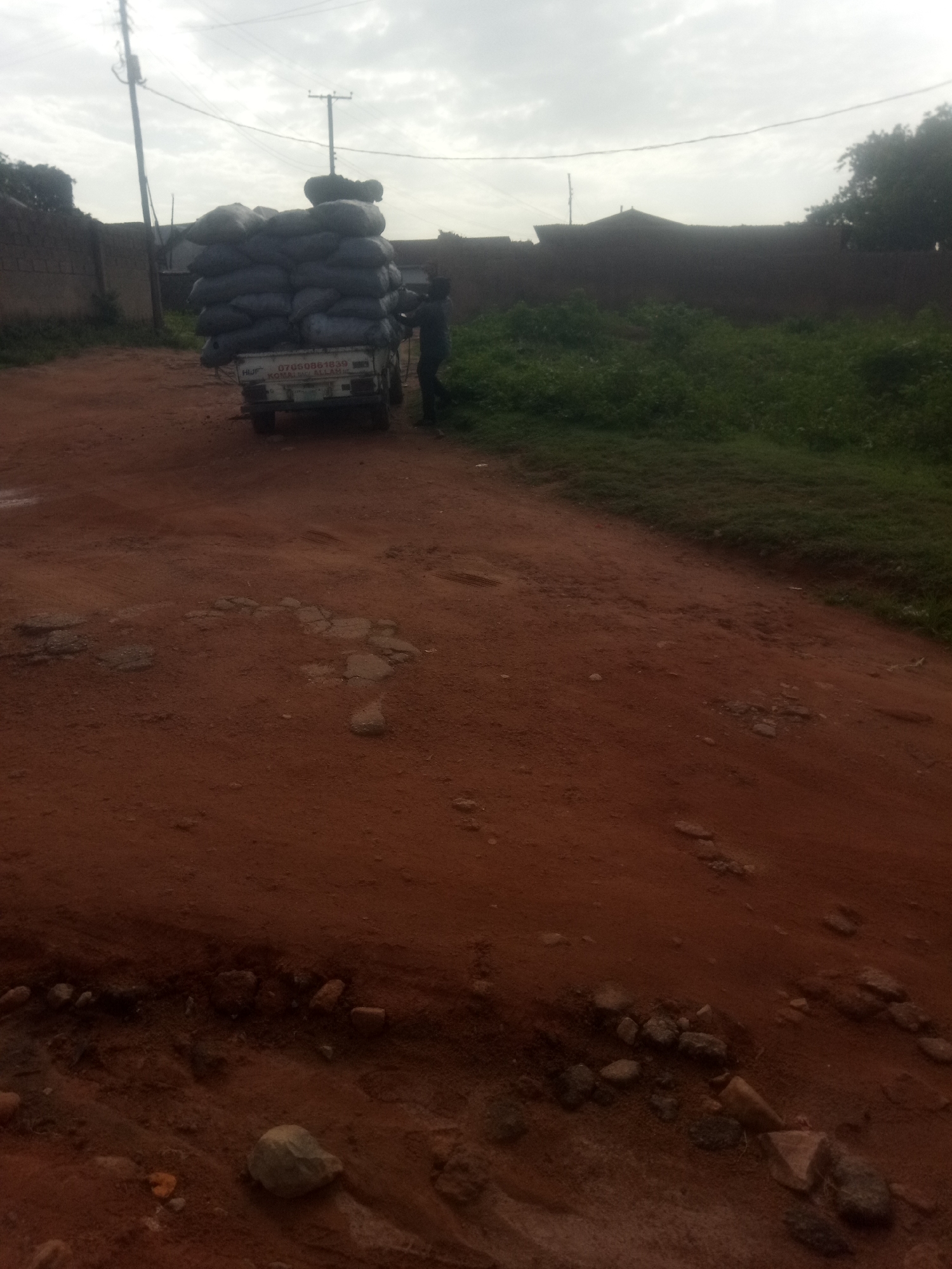 A man is seen standing next to a white truck loaded with bags of charcoal. The truck has license plates that read "HIJP" and "KOMALLAH". The man is wearing a dark shirt and pants. The truck is parked on a dirt road. The background shows a brick wall and some greenery. The sky is overcast. The scene is likely in a rural or semi-urban area.