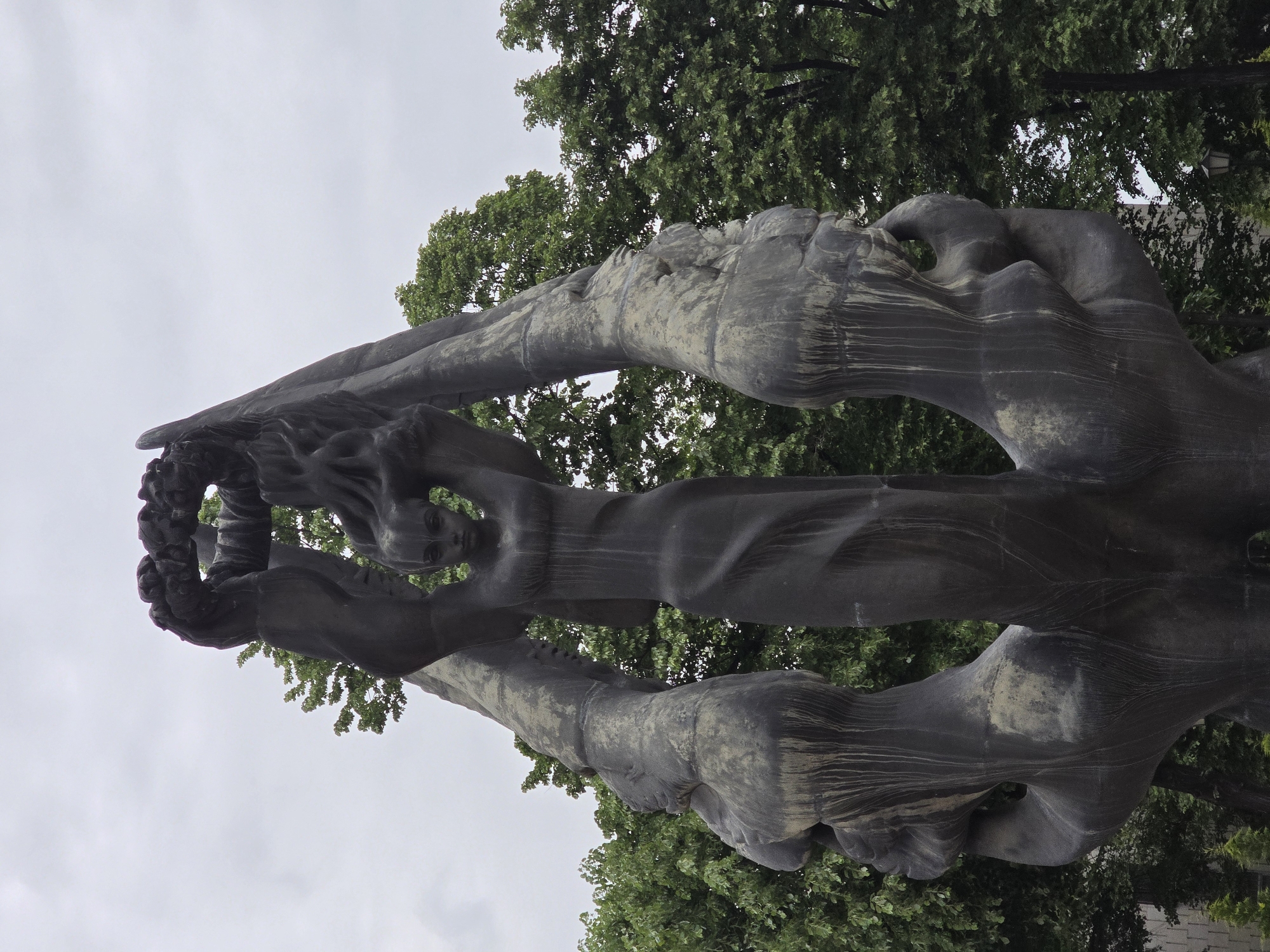 The image depicts a dark-colored sculpture of four figures intertwined, possibly made of stone or metal. The figures appear to be reaching upwards, with the uppermost figures holding what seems to be a wreath or circular object. The sculpture is set against a backdrop of green trees and a gray, overcast sky. The location is in the city of Unknown, Unknown.