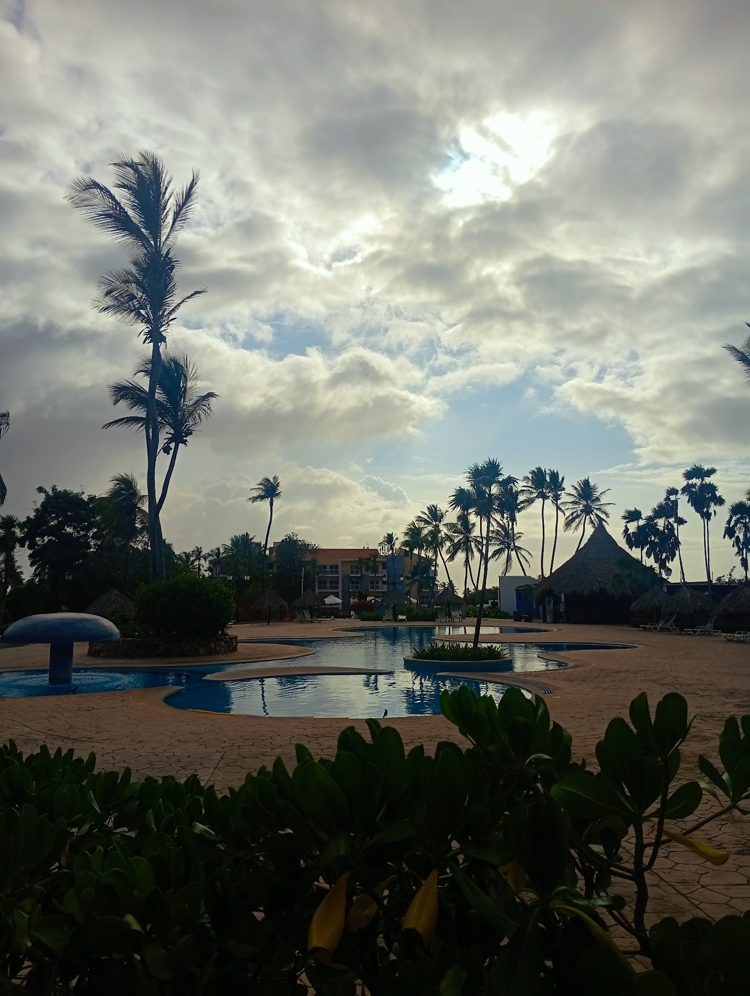 This image depicts a tranquil resort scene at Playa El Agua, Venezuela, under an overcast sky with the sun partially breaking through. The central focus is a large, irregularly shaped swimming pool complex filled with blue water, surrounded by light-colored paved decking.
In the foreground, dark green leafy bushes provide a natural frame for the lower portion of the image. To the left, a distinctive blue, mushroom-shaped water feature stands in one of the pool sections. Numerous tall palm trees, with their fronds appearing dark against the brighter sky, are scattered throughout the landscape, creating a tropical ambiance.
In the midground, a traditional thatched-roof hut (palapa) is visible to the right of the pool, likely serving as a bar or shaded relaxation spot. Further back, a multi-story hotel or resort building with a reddish-orange roof and visible balconies is nestled amongst the dense tropical foliage. The sky is dominated by thick, grey clouds, but patches of bright light indicate the sun breaking through, illuminating parts of the clouds and creating a diffused, somewhat dramatic light across the scene. No people are visible, lending a peaceful and serene atmosphere to the resort setting.