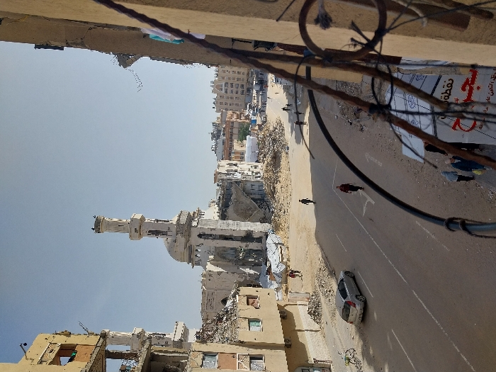 The image presents a high-angle view of a street scene in Khan Yunis, Palestinian Territory, under a clear, light blue sky during daylight. The central feature is a heavily damaged, light-colored mosque with a partially intact dome and a tall, structurally compromised minaret, which has birds or objects perched at its apex. Substantial rubble surrounds the mosque's base. To the left of the mosque, a multi-story building is extensively damaged, with exposed internal structures and debris.
A two-lane paved road, marked with white double solid lines and directional arrows, traverses the scene. Its unpaved shoulders are dusty and littered with rubble. A silver-grey sedan is parked on the left side of the road, near a bicycle leaning against a wall. Six individuals are visible: one person in a dark red top walks in the left lane; another in dark clothing walks further down the right side; two individuals are on the unpaved area to the left of the road; and two more, one wearing a light blue head covering, are partially visible in the lower right foreground near a white banner displaying Arabic text. The foreground includes a metal railing and the damaged facade of a building, from which light-colored clothes are hanging on the far right.