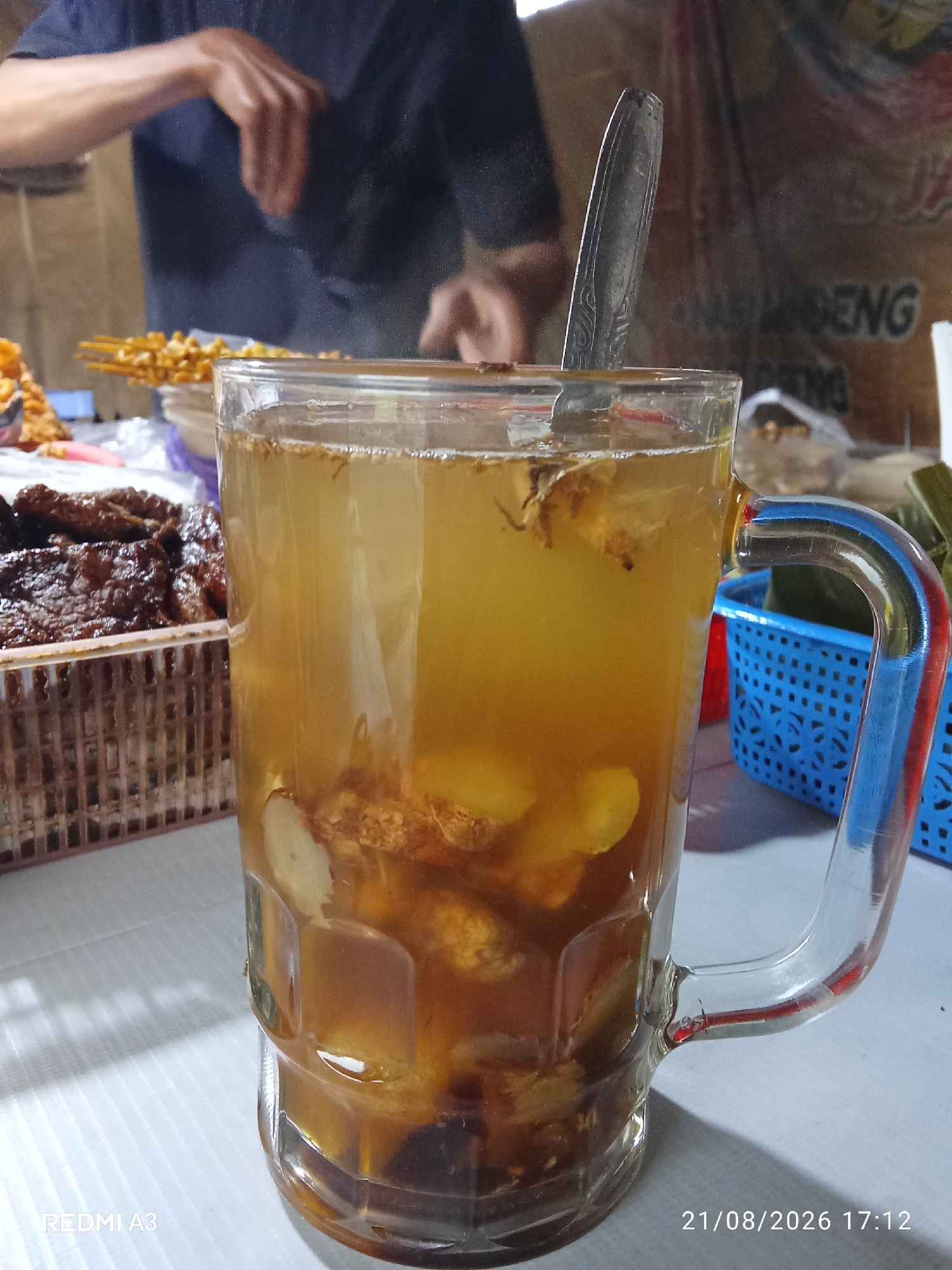 In Purbayan, Indonesia, a close-up shot captures a mug filled with a golden-brown herbal drink, featuring visible chunks of ginger and other roots. A metal spoon rests inside the mug, its handle extending slightly above the liquid. To the left, a basket overflows with dark, savory-looking meat, possibly jerky or a similar preparation, arranged in neat rows. In the background, a person wearing a dark blue shirt is partially visible, their hands engaged in an unseen task. Further back, a stall displays skewers of golden-brown food items, and a blue plastic basket holds green, leaf-wrapped packages. The scene is set on a white tablecloth, illuminated by ambient lighting. A timestamp in the lower right corner reads "21/08/2026 17:12".