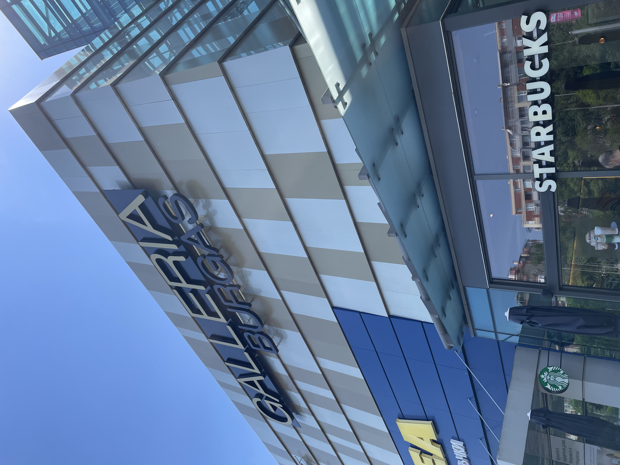 An upward-angled shot captures the exterior of a modern commercial building, identified as "GALLERIA BURGAS," under a clear blue sky. The facade features a striking pattern of alternating light and darker gray/beige horizontal panels, complemented by extensive glass elements, including what appears to be a glass canopy or roof.
Prominently displayed on the building's side are the large blue letters spelling "GALLERIA BURGAS." To the right, a "STARBUCKS" sign in white vertical lettering is visible on a glass window, which reflects nearby buildings and the bright sky. Below this, the iconic green Starbucks siren logo is seen alongside two dark, folded outdoor umbrellas. A partial "IKEA" logo, yellow on a blue background, is visible at the bottom of the building. Below the IKEA logo, a sign with Bulgarian text reads: "Кухнята е сърцето на дома. Направете я специална." (meaning "The kitchen is the heart of the home. Make it special."). The bright sunlight indicates it is daytime, with clear weather. The visible text confirms the location as Burgas, Bulgaria.
