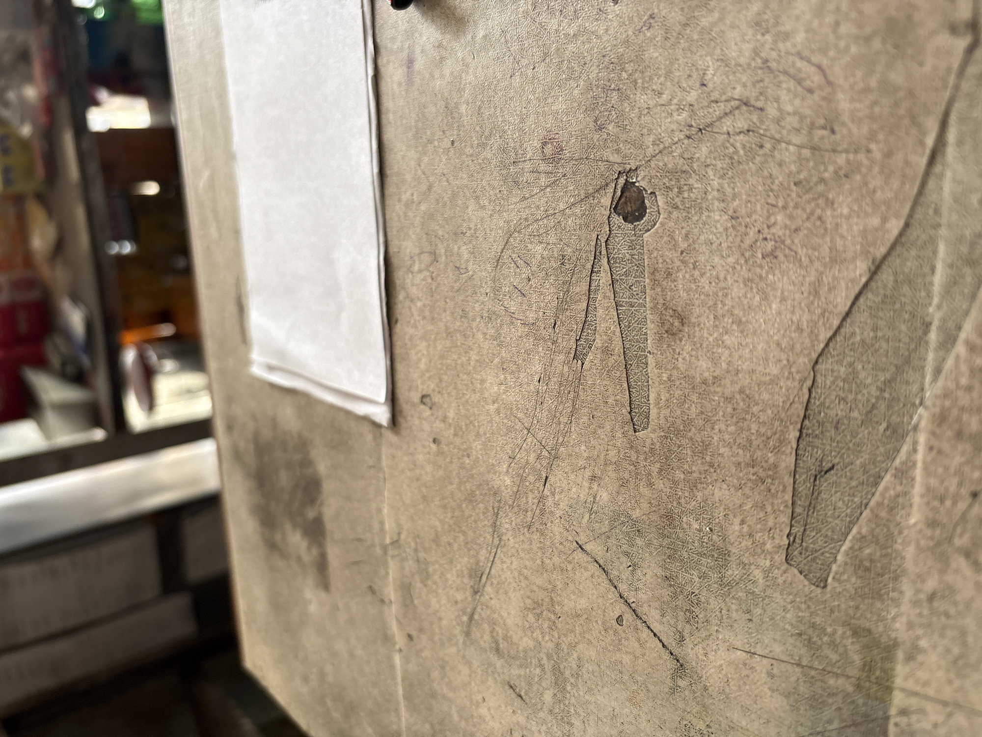 A close-up, slightly angled shot of a worn, light brown textured surface, possibly a table, reveals signs of age and use. Scattered markings, including faint purple lines that appear to be pen or pencil, are visible across the surface. In the upper right quadrant, a white sheet of paper is laid flat, partially covering the textured surface. The background is out of focus, showing shelves with various items, suggesting an indoor environment such as a shop or storage area. The lighting is somewhat dim, with light sources creating subtle highlights and shadows on the textured surface. There are no people visible in the frame. The image does not provide any specific details about Palimanan, Indonesia.