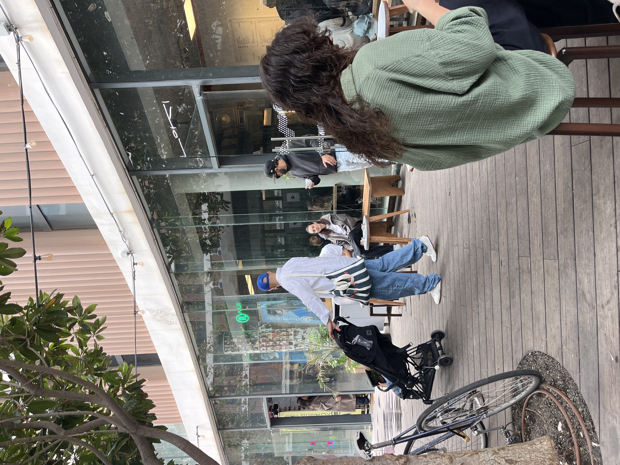The image shows an outdoor cafe setting in Tel Aviv, Israel. A woman with long curly hair wearing a green top sits at a wooden table. Other people are present at the cafe: a man in a black hoodie holds a cup, and a person in a beige coat is also visible. A man in a blue baseball cap and white shirt stands with a baby stroller. The cafe has a large glass facade, and some text is visible on the glass. A bicycle leans against a tree near the cafe's wooden deck. The outdoor area appears well-lit, suggesting it is daytime.