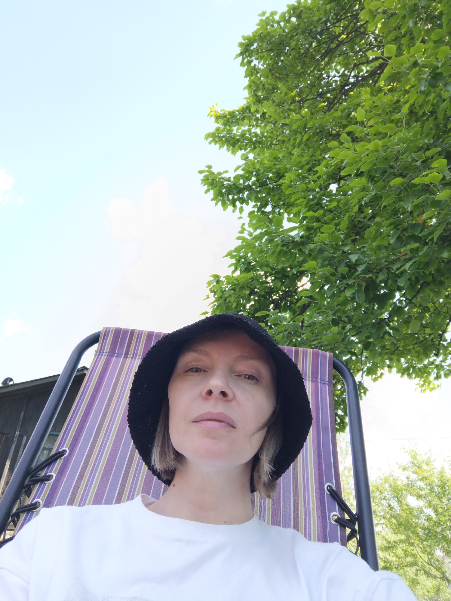 The image shows a person wearing a white shirt and a black bucket hat, seated in a purple-striped chair. Above and to the right of the person is a tree with dense green foliage, set against a light blue sky with faint white clouds. Parts of a building and other trees can be seen in the background. The location is likely Chaikovsky, Russia.