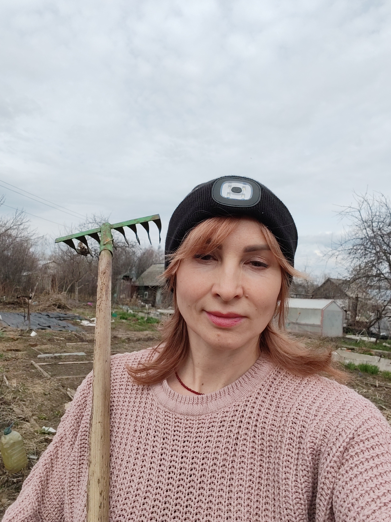 A woman stands in a garden in what appears to be a residential area. She is holding a rake and wearing a pink sweater and a black beanie with a light on the front. The background shows a cloudy sky, bare trees, a small greenhouse, and other buildings. There's a plastic jug on the ground to the left. The location is Chaikovsky, Russia.