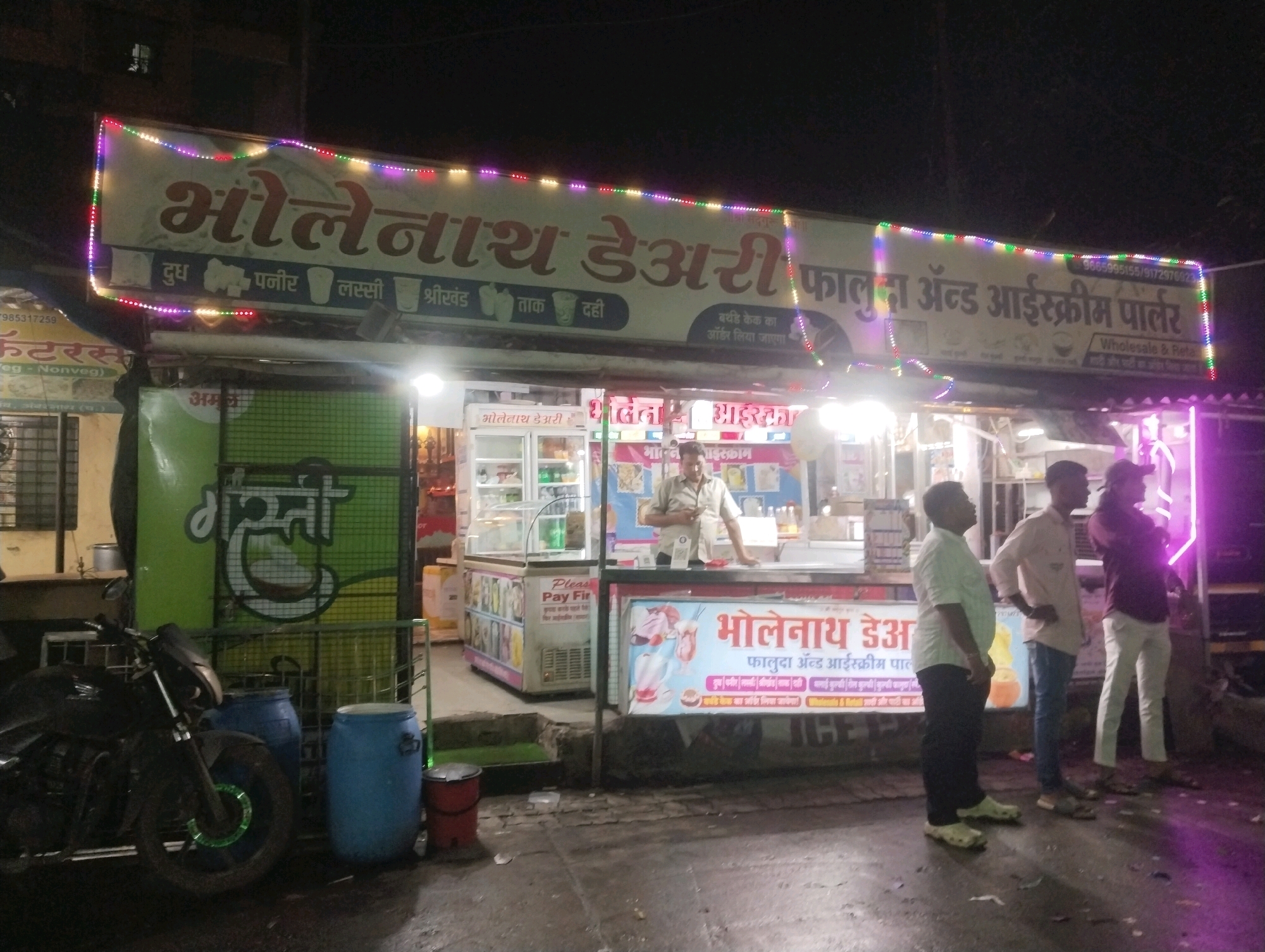 A night-time exterior view shows a roadside dairy and ice cream parlor in Ambernath, India. A large illuminated sign reads "Bholenath Dairy Falooda & Ice Cream Parlor" in Hindi script, with smaller text indicating "Wholesale & Retail." Beneath the main sign, more Hindi text lists products like "Dudh" (milk), "Paneer" (cheese), "Lassi" (yogurt drink), "Shrikhand" (sweetened strained yogurt), "Taak" (buttermilk), and "Dahi" (curd). Below the sign, a display cooler with various ice cream flavors is visible, with a person standing behind the counter. To the left, a motorcycle is parked next to two large blue barrels. The ground is wet, suggesting recent rain. Three men are standing in front of the parlor, appearing to be customers or employees. The overall scene depicts a typical Indian street-side business operating at night.