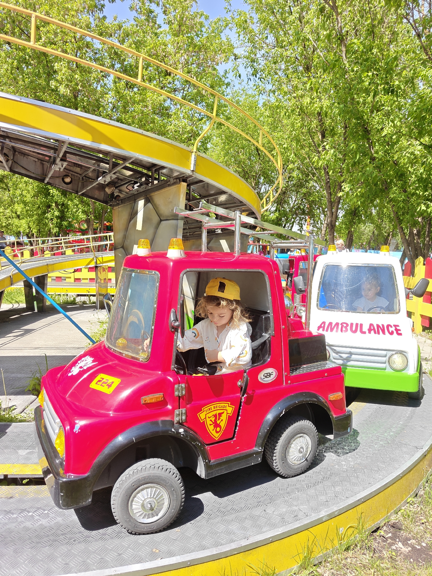 In Khimki, Russia, a young child with curly hair and a yellow hat is sitting in a red toy fire truck on a carousel-like ride. The fire truck has markings "224" and "FIRE BRIGADE." An ambulance toy vehicle is behind the fire truck, also occupied by a child. The ride is set outdoors on a sunny day with green trees in the background. A yellow elevated track with a metal base and a stone-like facade is also visible.
