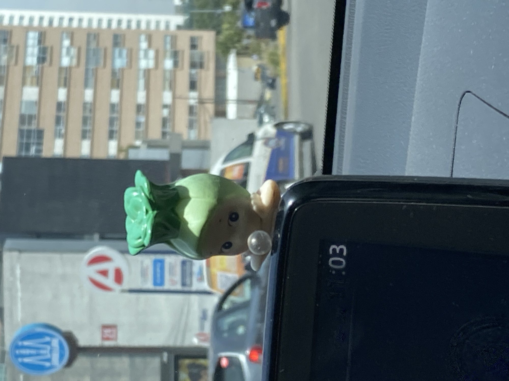 The image shows a Kewpie doll with a green vegetable hat stuck to the dashboard of a car, partially obscuring a GPS device displaying the time "11:03". Outside the car window, a building with numerous windows is visible, along with a street scene that includes a car and signs with a red "A" logo and a blue logo that says "WLV". The location is presumed to be a city, but without additional details, it cannot be identified.