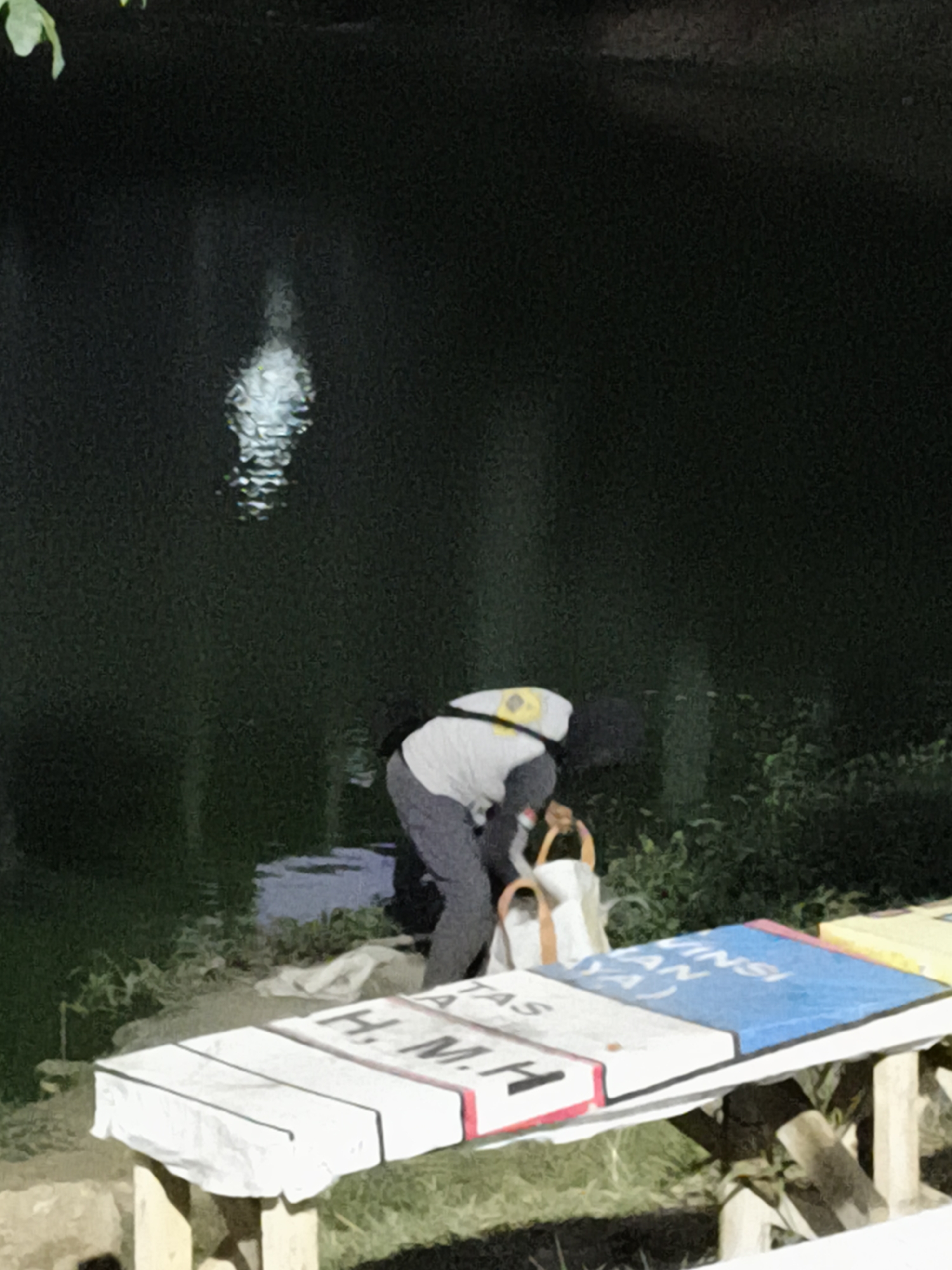 In Surabaya, Indonesia, a person is visible near the edge of a dark body of water, possibly a river or lake. The individual is bending over, appearing to be interacting with objects on the bank. They are wearing a light-colored long-sleeved shirt and dark pants. A yellow cap or hat with a reflective element is on their head, and they are holding a white bag with orange handles. Beside the water, the ground is covered with vegetation and some white cloth or material. In the foreground, a bench is set up, covered with what appear to be large, book-like objects. These objects are decorated with text and colors, including blue and white with red trim on one, and white with red and black markings on another. The scene is dimly lit, suggesting it is either dusk or nighttime, with a reflection visible on the surface of the water.