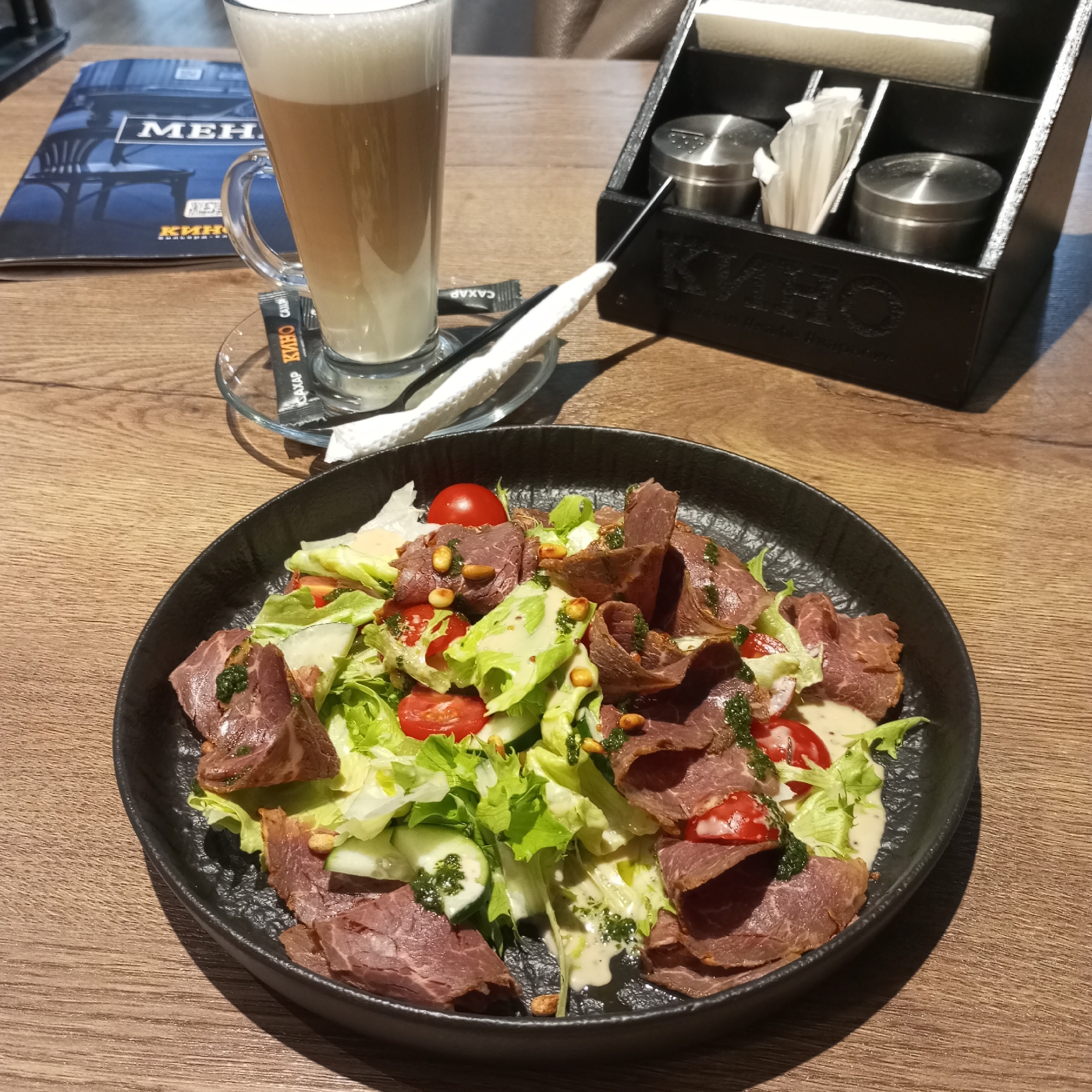 A close-up, eye-level shot shows a plate of salad and a tall glass of latte on a wooden table, likely in a restaurant or cafe. The salad, served on a dark, textured plate, contains sliced meat, lettuce, cherry tomatoes, cucumber slices, and pine nuts, all dressed in a creamy sauce and drizzled with green pesto. To the left of the salad is a tall glass of latte with a thick layer of foam, placed on a saucer. Next to the glass is a sugar packet with Cyrillic writing. In the background, a dark wooden condiment caddy holds salt and pepper shakers, napkins, and toothpicks. A menu with the word "МЕНЮ" (Menu in Russian) is visible on the left side of the frame. The lighting suggests an indoor setting, possibly natural light filtering in. The establishment is likely located in Izhevsk, Russia, given the Cyrillic text on the menu and sugar packet.