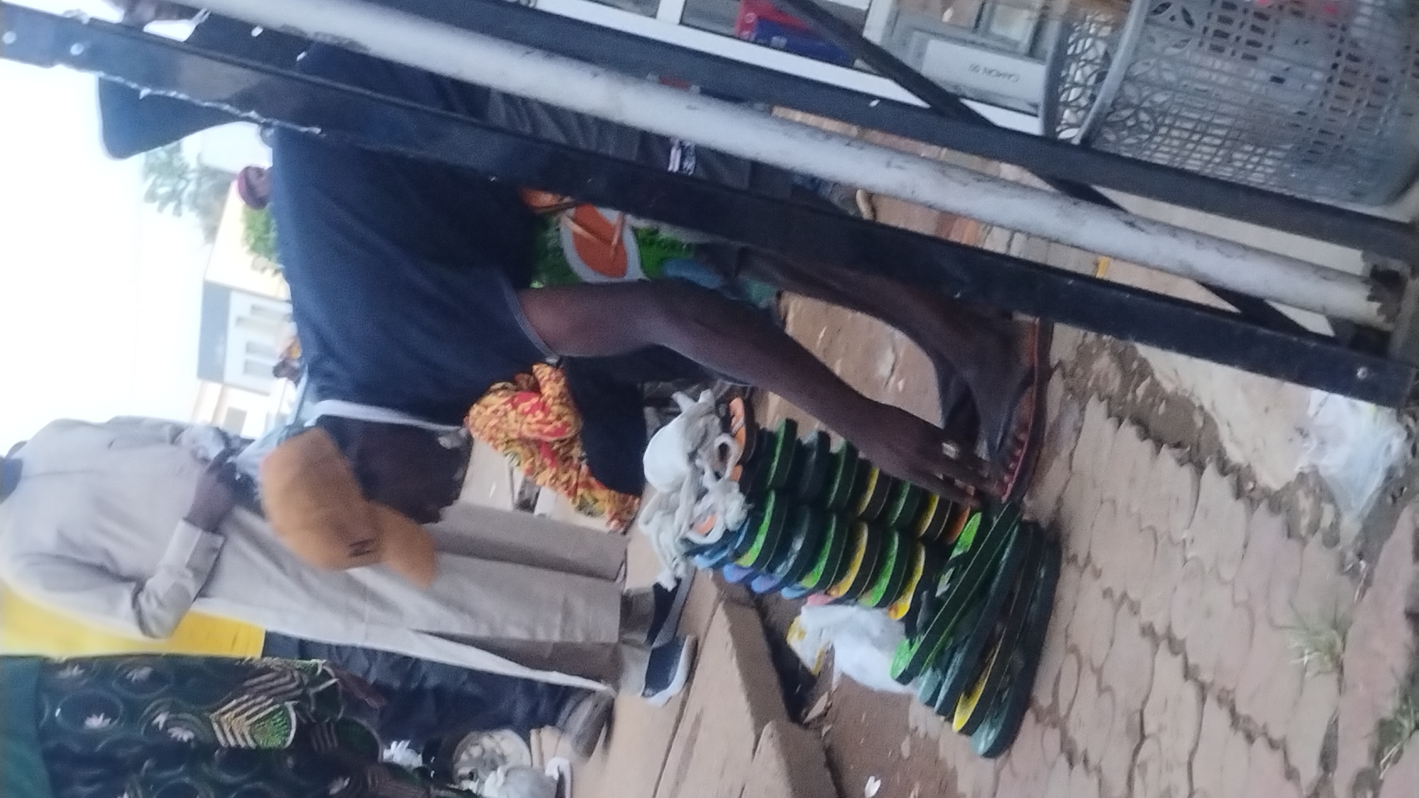The image captures an outdoor market scene, taken from a slightly high and tilted angle. In the foreground, a person wearing a dark blue shirt and colorful shorts or fabric is bent over, seemingly arranging or tending to a large collection of flip-flops. These flip-flops, primarily in shades of green and yellow, are neatly stacked or laid out on the ground, possibly on a mat or tarp. Near the bending person, some white bundled items, resembling fabric or more footwear, are also visible.
To the left, another person stands, dressed in a light-colored (likely white or beige) long-sleeved garment and wearing a yellow cap. Their hands are clasped or holding an object near their waist. The ground beneath the market stall is paved with interlocking bricks or tiles. Various other objects are scattered around, including patterned fabrics (one red/orange/yellow, another green/black) and a pair of dark blue sneakers.
In the background, metal frames and poles suggest a market stall structure, and a grey plastic crate or basket is perched on one of these structures. Faintly visible text on a white surface in the upper background reads "CANON" and "SG 90" or "SG 96". The scene appears to take place during the daytime, with sufficient ambient light for outdoor selling. The overall impression is that of a bustling, informal market.