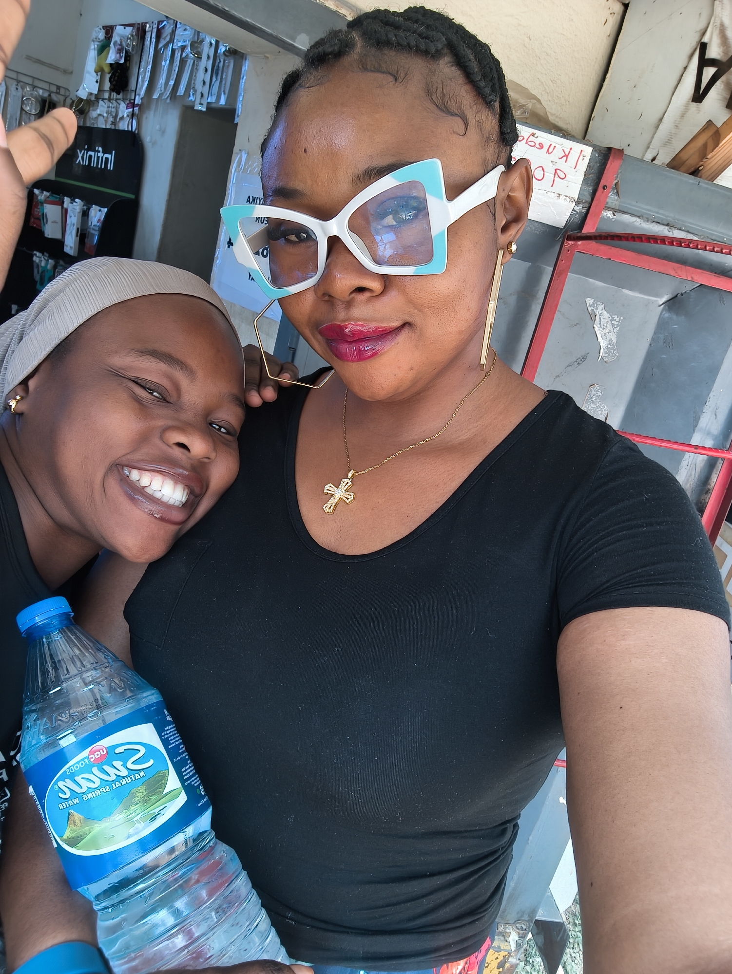 A vibrant, brightly lit selfie captures two Black women posing cheerfully in what appears to be a small shop or market stall in Jos, Nigeria. The woman on the right, positioned in the foreground, takes the photo with an outstretched arm. She wears a black t-shirt, striking white and light blue geometric sunglasses, long golden hexagonal hoop earrings, and a gold necklace featuring a cross pendant. Her hair is styled in braids, and she sports bright red lipstick. Leaning into her on the left is another woman, smiling broadly with white teeth. She wears a light grey head covering or turban and a black top, and holds a blue plastic bottle of "SWAN NATURAL SPRING WATER" (visible in inverted text).
The background suggests a commercial setting, possibly displaying merchandise on shelves or racks, with a dark green banner showing partially inverted text, likely "Infinix" (as "xinitnl"). Other fragmented and inverted text, such as "AXIYA" and partial words like "PO?" on various signs, are also visible. The lighting indicates it is daytime. Both women are smiling, conveying a sense of happiness and camaraderie.