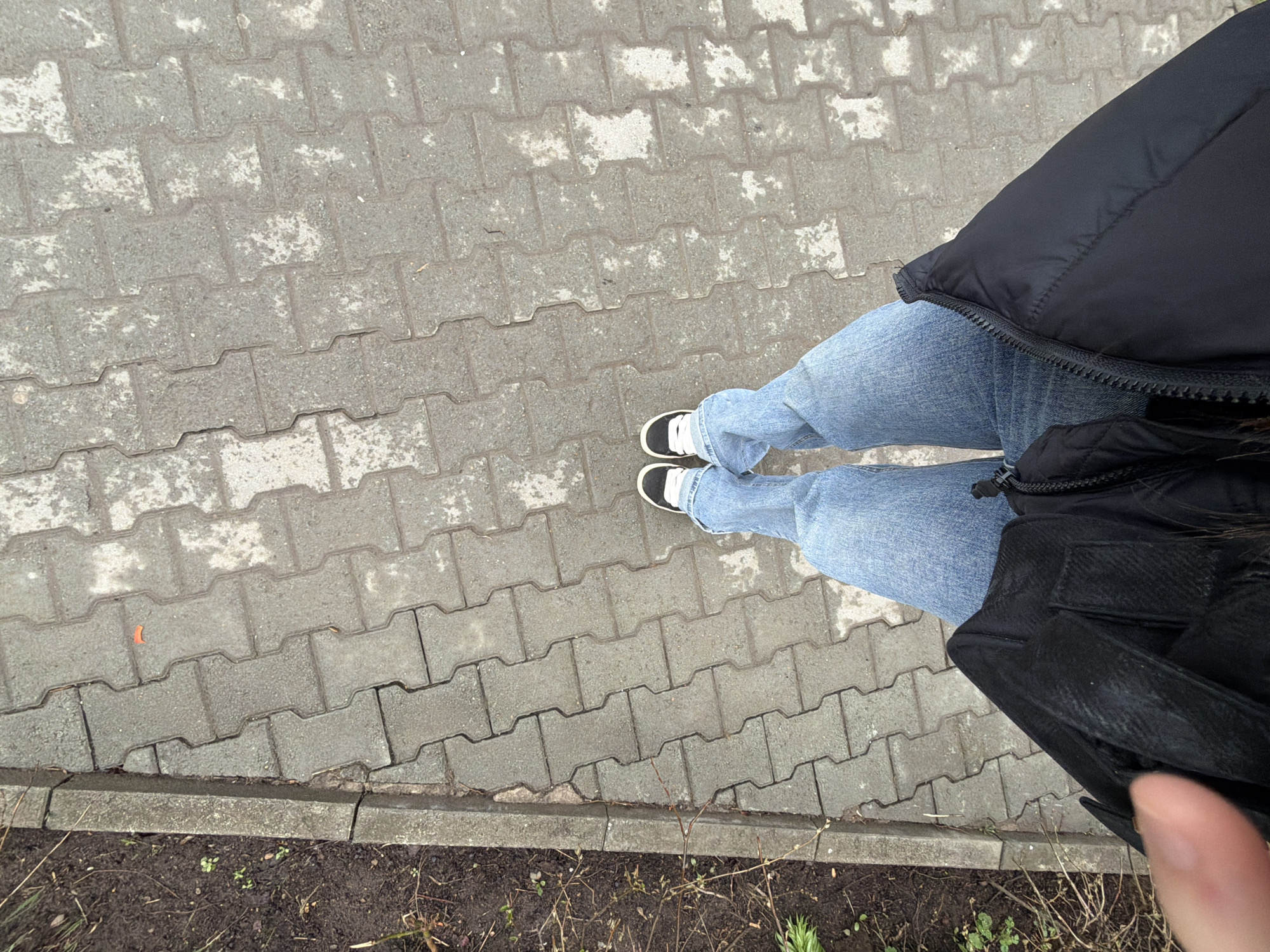 This top-down shot captures a person standing on a grey paved surface in Chaikovsky, Russia. The individual is wearing light blue jeans, black and white sneakers with white laces, and a black puffer jacket. A partial view of their right hand, specifically a thumb, is visible in the bottom right corner.
The scene features an outdoor pathway constructed from interlocking grey concrete paving stones, many of which show darker, mottled areas, suggesting they might be damp or aged. A concrete curb separates the paved path from a narrow strip of dark, loose soil. This soil patch contains sparse vegetation, including dry, thin branches and a few small green shoots. The lighting is dull and even, indicating an overcast day. No text is visible in the image. The activity depicted is simply standing or momentarily paused on the pathway.