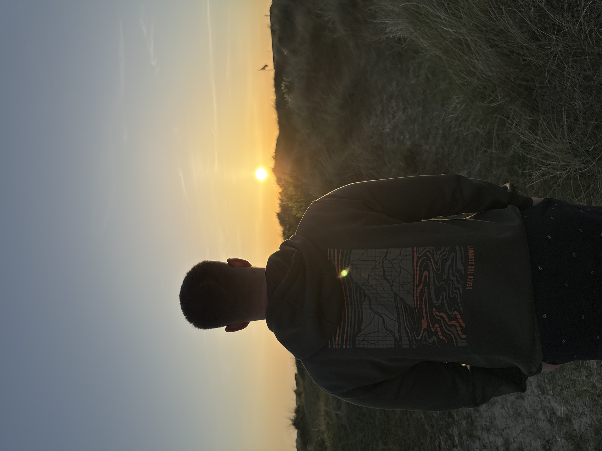 A medium shot depicts a person from behind, silhouetted against a sunset. The person is wearing a green hoodie with a design on the back and dark shorts. The setting appears to be a grassy dune area in Great Yarmouth, United Kingdom. The sun is setting on the horizon, creating a golden glow in the sky. A bird is visible in the sky above the sun. The environment includes dry grasses in the foreground and background. The person is stationary, seemingly observing the sunset.