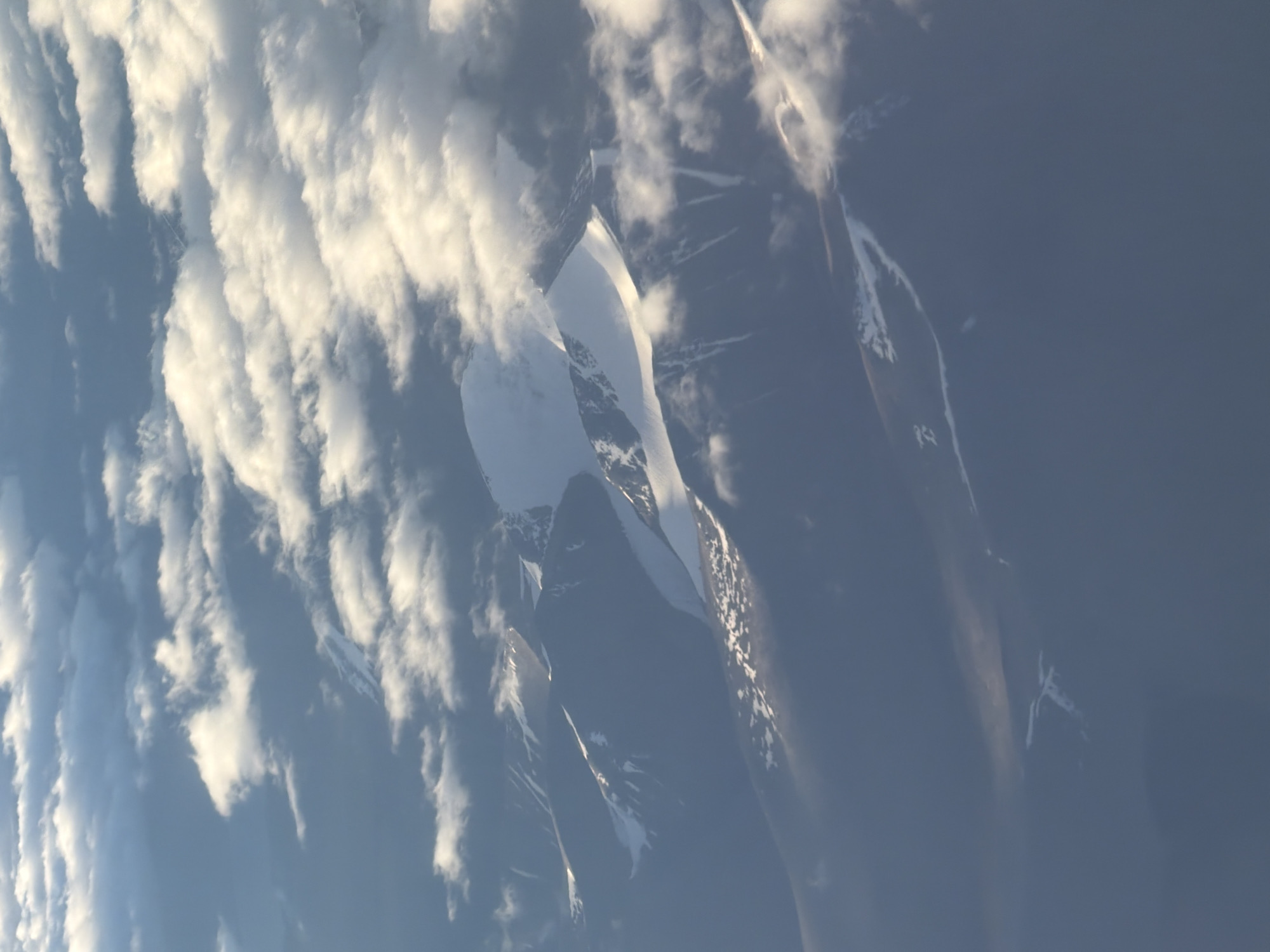 An aerial view captures a mountainous landscape covered in snow and clouds. The peaks are rugged and sharp, with patches of white snow contrasting against the darker rock. Clouds drift over and around the mountains, some obscuring the view, while others are illuminated by sunlight. The scene suggests a high-altitude environment, possibly during winter or in a region with permanent snow cover. The image does not depict the city of Hiranagar, India.