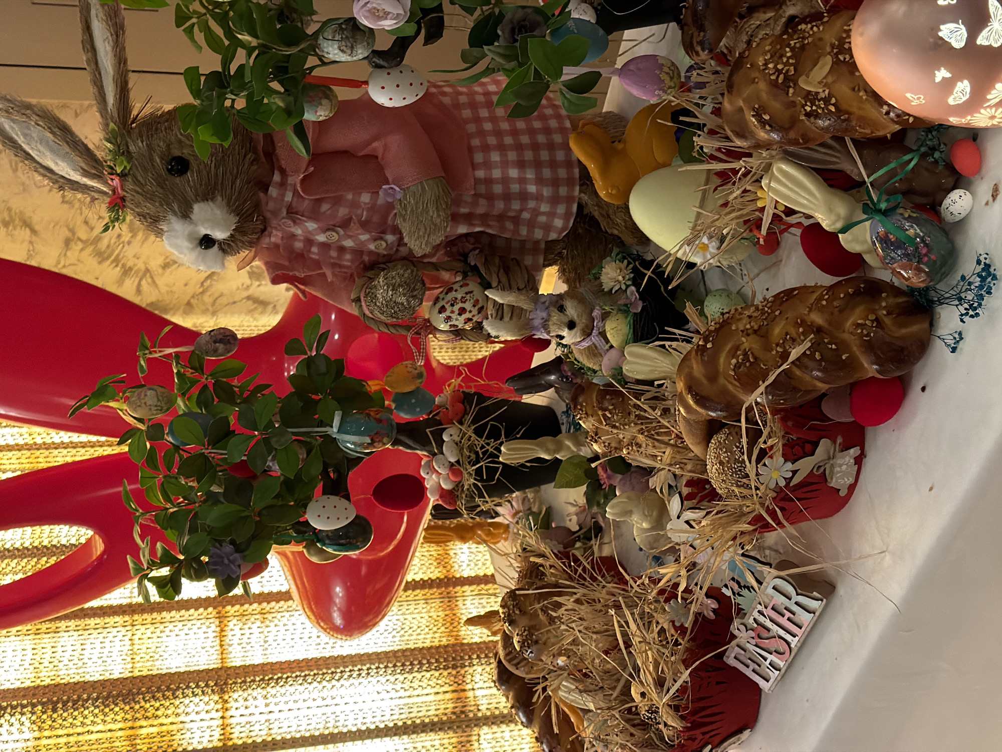 This image appears to be a close-up photograph of an Easter display, possibly indoors.
**Subjects and Objects:** The display features a straw rabbit wearing a pink gingham dress, surrounded by Easter-themed decorations. These decorations include painted Easter eggs, possibly homemade, decorative flowers and foliage, and several braided loaves of bread topped with nuts and seeds. A pink decorative light with butterflies is also present. There are also decorative Easter bunnies. A red vase, shaped like a bow, is present on the display. A wooden sign with the word "Easter" is visible. The background appears to be window blinds.
**Environment:** The setting appears to be inside, likely a home or shop, given the presence of the display. The background suggests that there may be a window with blinds behind the display. There is no information available in the image about the city of Budva, Montenegro.