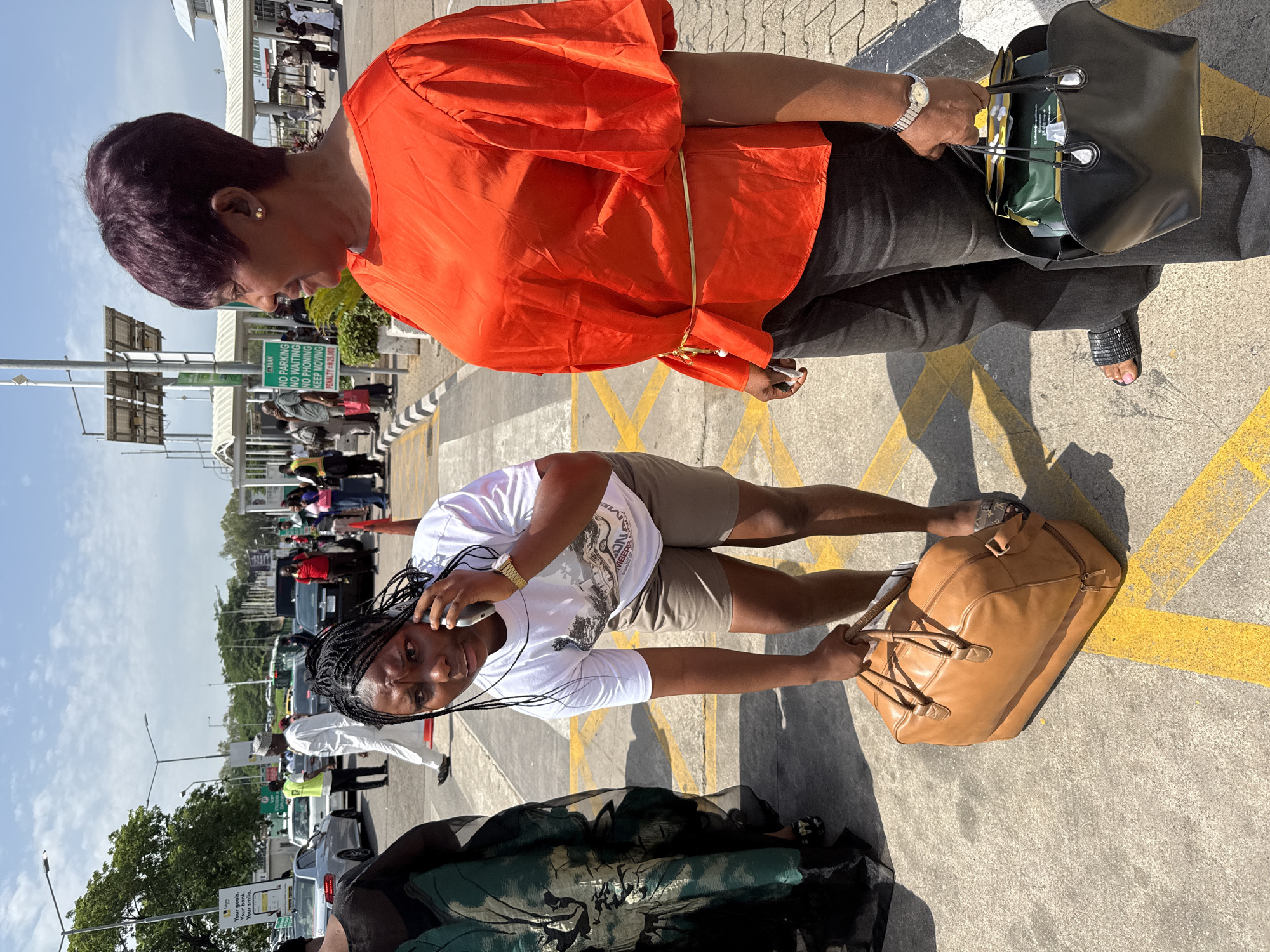 In Gwaska, Nigeria, a young woman with braided hair is on the phone, bending over to pick up a brown leather duffel bag. She is wearing a white t-shirt and khaki shorts. To her right stands another woman with short, dark hair, wearing a bright orange top and black pants. She is holding a black handbag filled with various items, including what appears to be a green box or package. They are standing on a paved area with yellow markings, likely an airport tarmac or parking lot, under a bright, sunny sky with some clouds. In the background, there are several vehicles, including cars and what might be a bus, as well as other people and airport infrastructure. A sign with "NO PARKING NO WAITING NO PHONING KEEP MOVING" is visible in the middle ground.