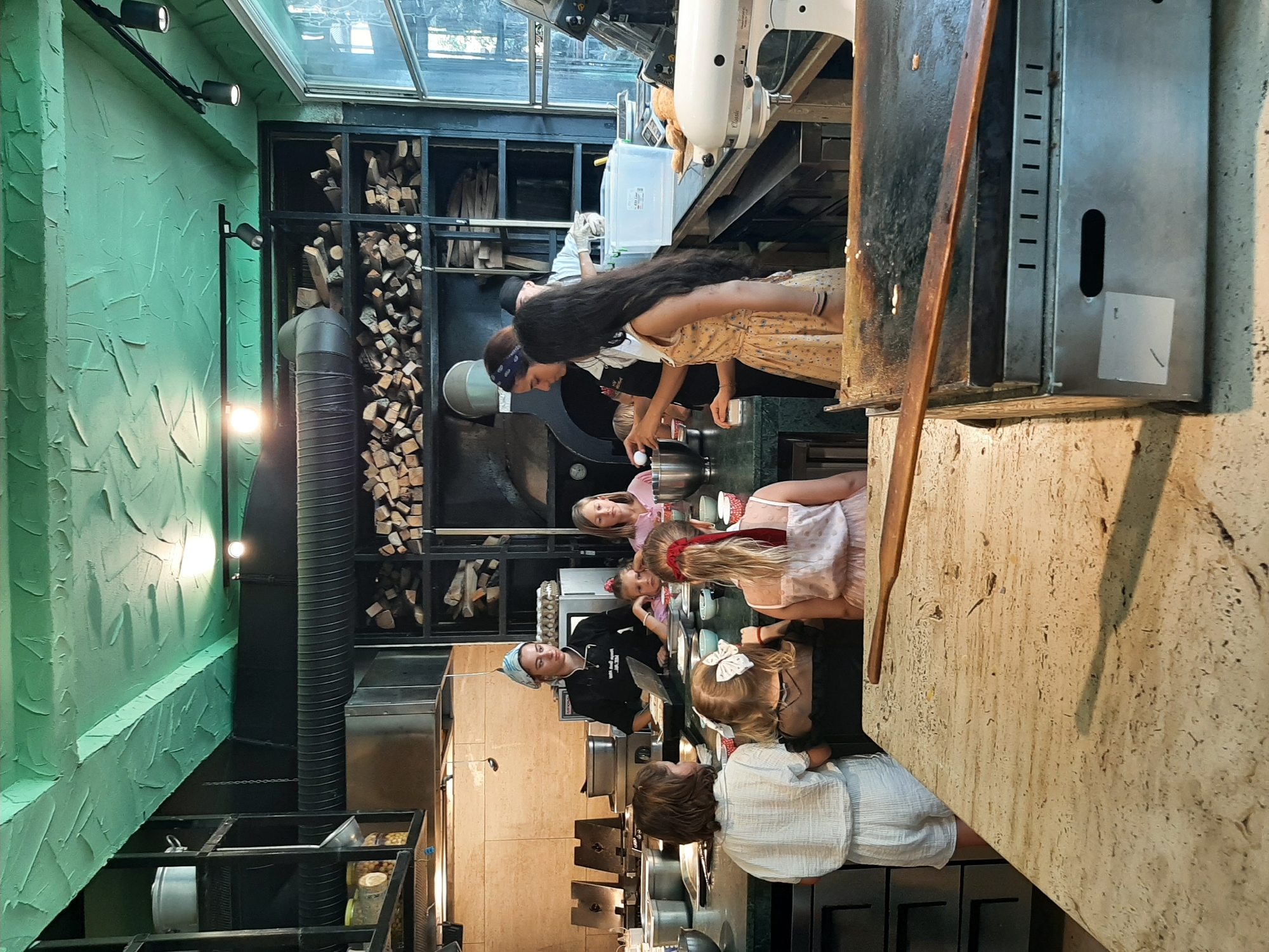 A group of people are gathered around a kitchen counter in a restaurant. There are four young girls, one woman in a chef's uniform, and two women in casual clothing. The girls are facing the counter, and two of the women are standing behind it, appearing to be instructing or assisting them. One of the girls is wearing a chef's hat. Various kitchen utensils and equipment are visible on the counter, including bowls, whisks, and a mixer. In the background, there are shelves filled with logs and cooking appliances. The ceiling is painted a textured green, and there is a large window on the right side of the frame. The overall scene suggests a cooking class or a culinary demonstration. The image does not contain information to determine the location within Alanya, Turkey.