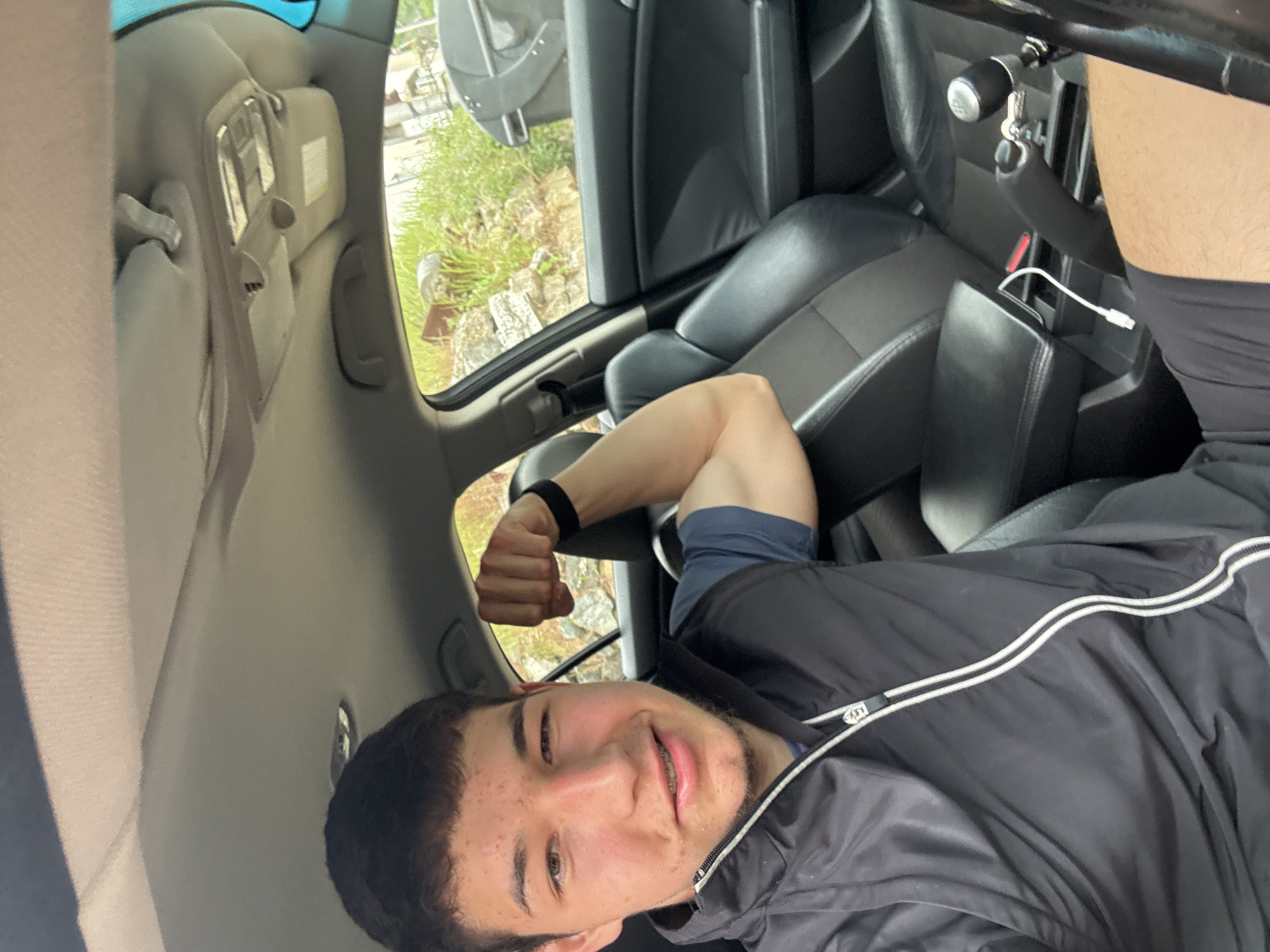 The image shows a person in the driver's seat of a car. The person is smiling at the camera and flexing their arm. The interior of the car is visible, including the seats, steering wheel, and dashboard. Outside the car, greenery can be seen through the window. There is no indication of Zlatograd, Bulgaria.