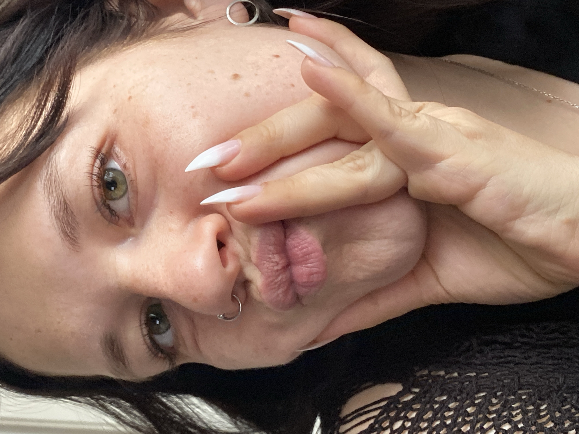 A close-up, tilted portrait shot of a light-skinned person with freckles. They have dark hair, green eyes, and are wearing a small silver nose ring and hoop earrings. Their lips are pursed, and one hand is positioned close to their face with long, white-tipped nails. The person is wearing a black, patterned top. The location is not identifiable from the image.