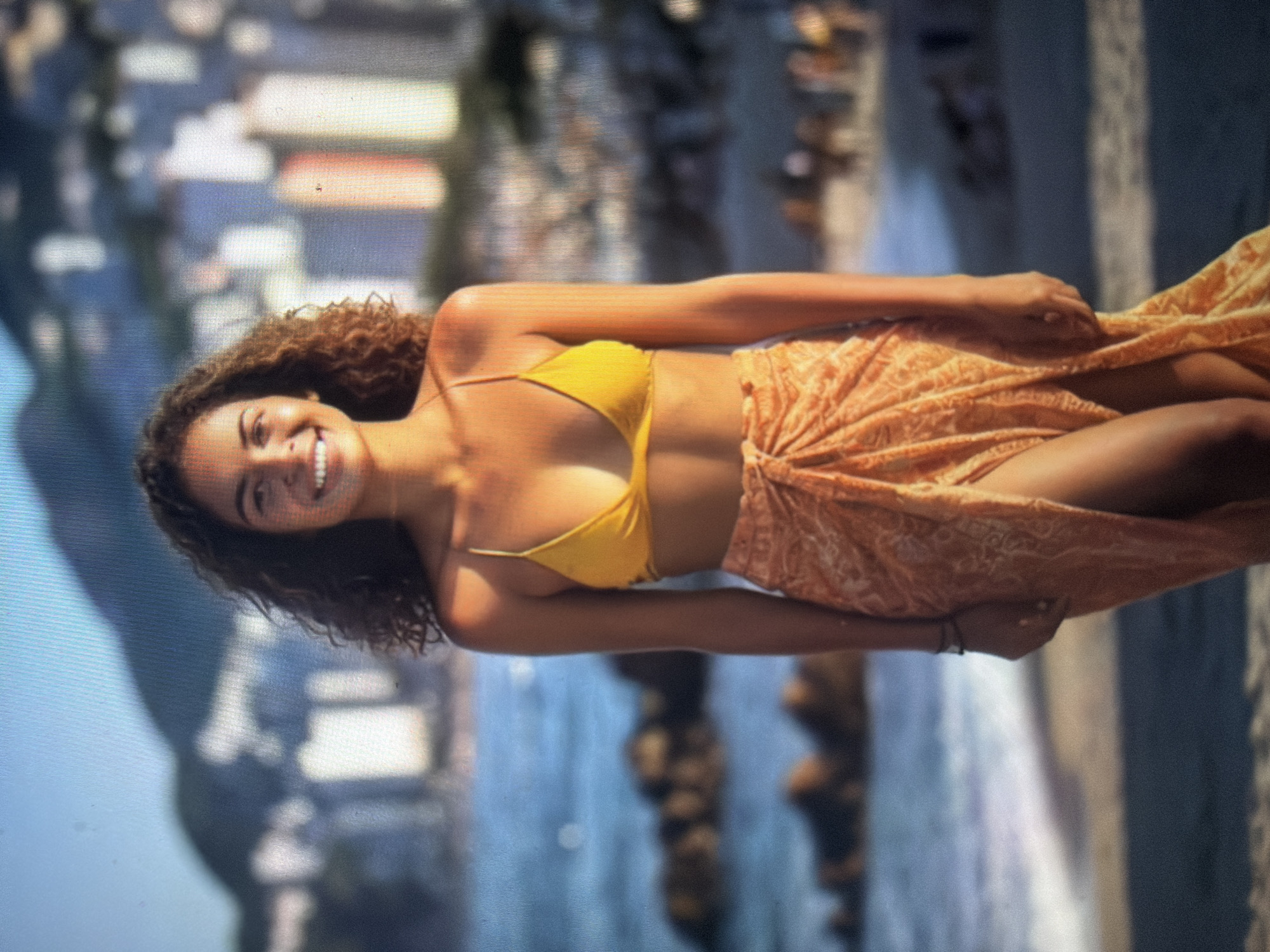 A young adult female is depicted standing centrally on a sandy beach. She has dark, curly hair and a direct gaze, exhibiting an open-mouthed smile that reveals her teeth. She wears a yellow string bikini top and an orange patterned sarong, which is wrapped around her lower body, revealing her right leg and a portion of her left arm. A thin, dark bracelet is visible on her left wrist. The immediate foreground shows a stretch of sand. Behind her, the ocean is visible with minor surface agitation, and several dark, rounded rock formations emerge from the water. The background presents a blurred urban landscape consisting of numerous multi-story buildings, appearing in shades of white, grey, and orange-brown. These structures are situated against a backdrop of large, dark mountains under a light blue sky. The scene is illuminated by natural daylight. This image is located in the city of Shumen, Bulgaria.