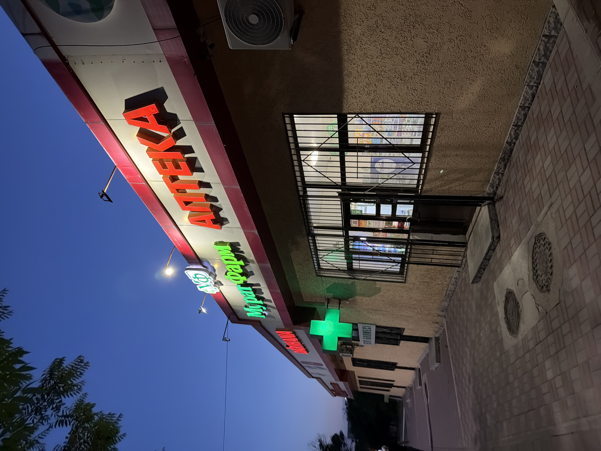 An exterior shot of a pharmacy at dusk shows a building with a stucco facade and a dark red trim above the windows and doors. The building's signage, illuminated in red and green neon, reads "АПТЕКА" (Apteka), which translates to "Pharmacy" in Russian, and "Мурат Фарм" (Murat Pharm). A bright green illuminated cross, a common symbol for pharmacies, is mounted on the facade. The building has large, barred windows and a doorway. An air conditioning unit is visible on the right side of the building. The ground in front of the building is paved with interlocking bricks. The sky is a deep blue, indicating the late hour. In the background, a tree with dark green leaves is silhouetted against the sky. This pharmacy is located in the city of Shymkent, Kazakhstan.