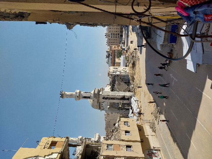 An elevated view captures a street scene in Khan Yunis, Palestinian Territory, marked by extensive structural damage. Dominating the mid-ground is a significantly damaged white mosque, featuring a tall, slender minaret with a multi-tiered top and a central dome, partially collapsed. Surrounding the mosque are piles of rubble. To the left, a multi-story, light brown building shows severe damage, with missing sections of its facade and exposed interiors. Similar destruction is evident in other buildings throughout the background. The paved street below is partially covered with dirt and debris, displaying white lane markings and a directional arrow. Approximately 15 individuals, predominantly young males, are scattered across the street; a group of about ten are actively playing, likely soccer, in the central roadway. Other individuals walk further down the street. On the immediate right, the edge of a building or balcony is visible, with a metal railing from which a piece of denim clothing and a pink clothespin hang. Small, makeshift stalls with blue and white coverings line the right side of the street near the foreground. The sky is clear blue. Overhead, thin wires span between buildings.