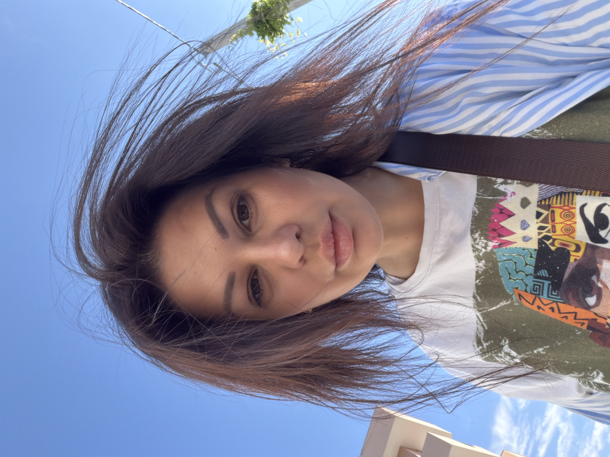 A female adult is depicted in a low-angle selfie, looking directly at the camera against a clear blue sky in Sochi, Russia. Her long, dark brown hair is disheveled and windblown, with numerous individual strands visible against the sky. She has brown eyes, a neutral expression, and full lips slightly parted. She is wearing a white t-shirt with a complex, multi-colored graphic print on the chest, layered under an open light blue and white striped shirt, visible on her right side. A dark brown strap, possibly from a shoulder bag, runs diagonally across her chest from her left shoulder. In the lower left background, a portion of a light-colored building with rectilinear features is visible. To the mid-right, a utility pole or lamppost partially covered in green foliage is present, with a thin, light-colored wire extending upwards into the sky. The scene is illuminated by bright daylight, indicating clear weather and a light breeze.