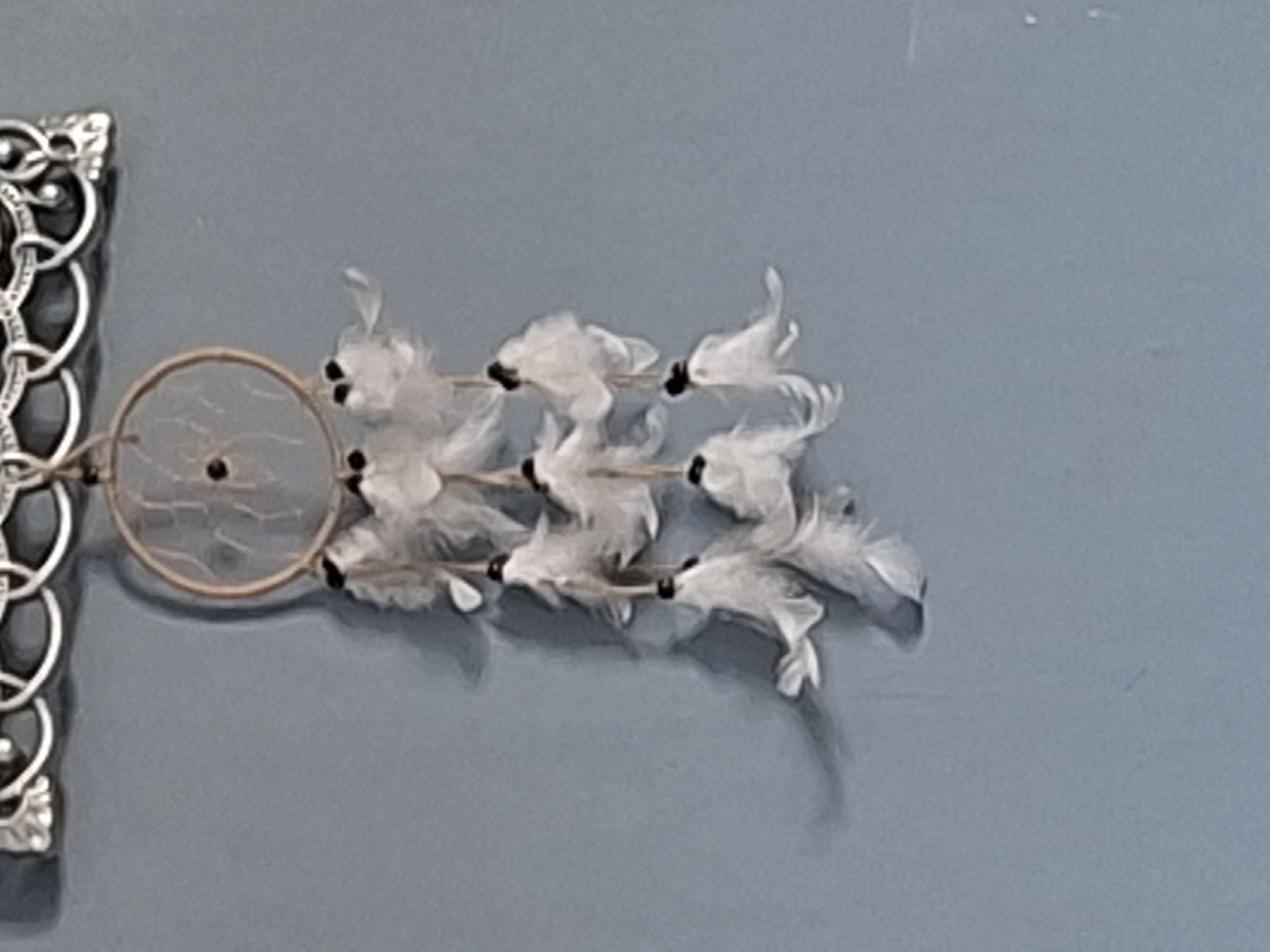 The image depicts a dreamcatcher hanging on a light blue wall. The dreamcatcher consists of a circular frame with a woven net inside and three rows of white feathers attached to it with black beads. To the left of the dreamcatcher is a decorative metal element with intricate patterns. The scene is indoors. The image does not provide enough information to determine the location or the city.