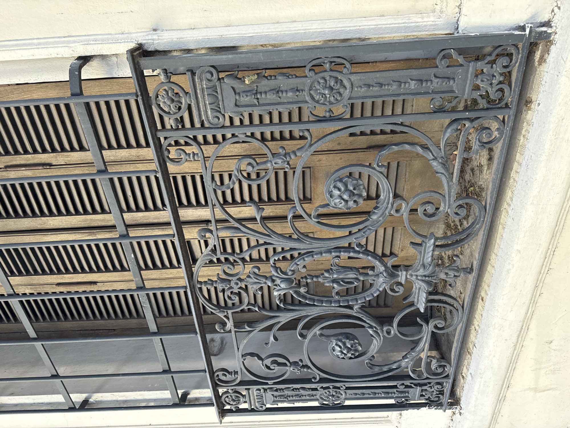 An upward-facing view of an ornate wrought iron balcony in Buenos Aires, Argentina. The balcony is predominantly gray-black with detailed scrollwork and floral accents. Behind the wrought iron is a wooden slatted window frame. The balcony is attached to a building with a light-colored, possibly white, exterior. The image is rotated clockwise, presenting an unconventional perspective of the structure.