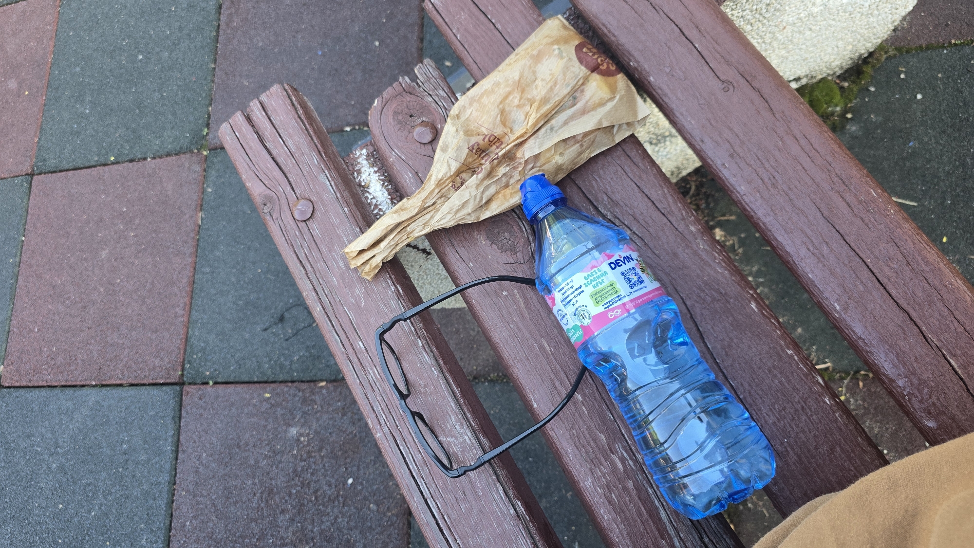 The scene is a detail shot of a brown, weathered wooden bench, situated outdoors, likely in a park or public space. The bench is made of planks with visible wood grain and has metal fasteners visible. Resting on the bench are several items. There is a crumpled brown paper bag, likely from a food purchase. A blue plastic water bottle with a blue cap, labeled "DEVIN" in Cyrillic script, is also present. Beside the water bottle, there is a pair of black eyeglasses with thin frames. The surface around the bench appears to be a playground or recreational area with alternating dark red and dark gray rubber tiles. A small patch of green moss is visible near the top right of the frame. A portion of a garment in a warm tan color appears in the bottom right corner of the image. There are small traces of white powder on the bench near the paper bag. This image was most likely taken in Sofia, Bulgaria due to the Cyrillic labels.