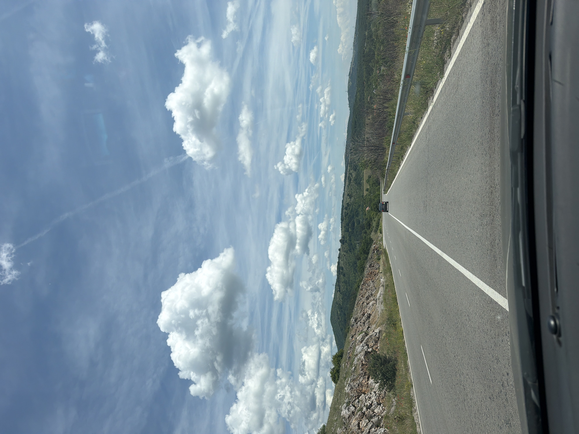 The image, taken from inside a vehicle looking forward, captures a scenic drive on a clear day in Bogyovtsi, Bulgaria. The perspective is from the passenger side, with part of the car's interior visible on the far right.
The central subject is a two-lane asphalt road, stretching into the distance with a solid white line marking the right edge and a broken white line down the center. A dark-colored car is visible ahead, further down the road. To the right of the road, a metal guardrail runs parallel to the asphalt, separating it from the shoulder.
On both sides of the road, the terrain is rugged and hilly, covered with sparse vegetation, rocks, and some patches of green grass. In the background, rolling mountains or large hills are visible, densely covered with trees and greenery, suggesting a rural or mountainous region.
The sky dominates the upper portion of the image, displaying a bright blue hue with numerous white, fluffy cumulus clouds scattered across it. There are also some fainter, wispy cloud formations. A faint contrail from an aircraft is visible in the upper left of the sky. The sunlight indicates it is daytime, likely mid-morning or afternoon, with fair weather conditions. No people are visible, and there is no discernible text in the image.