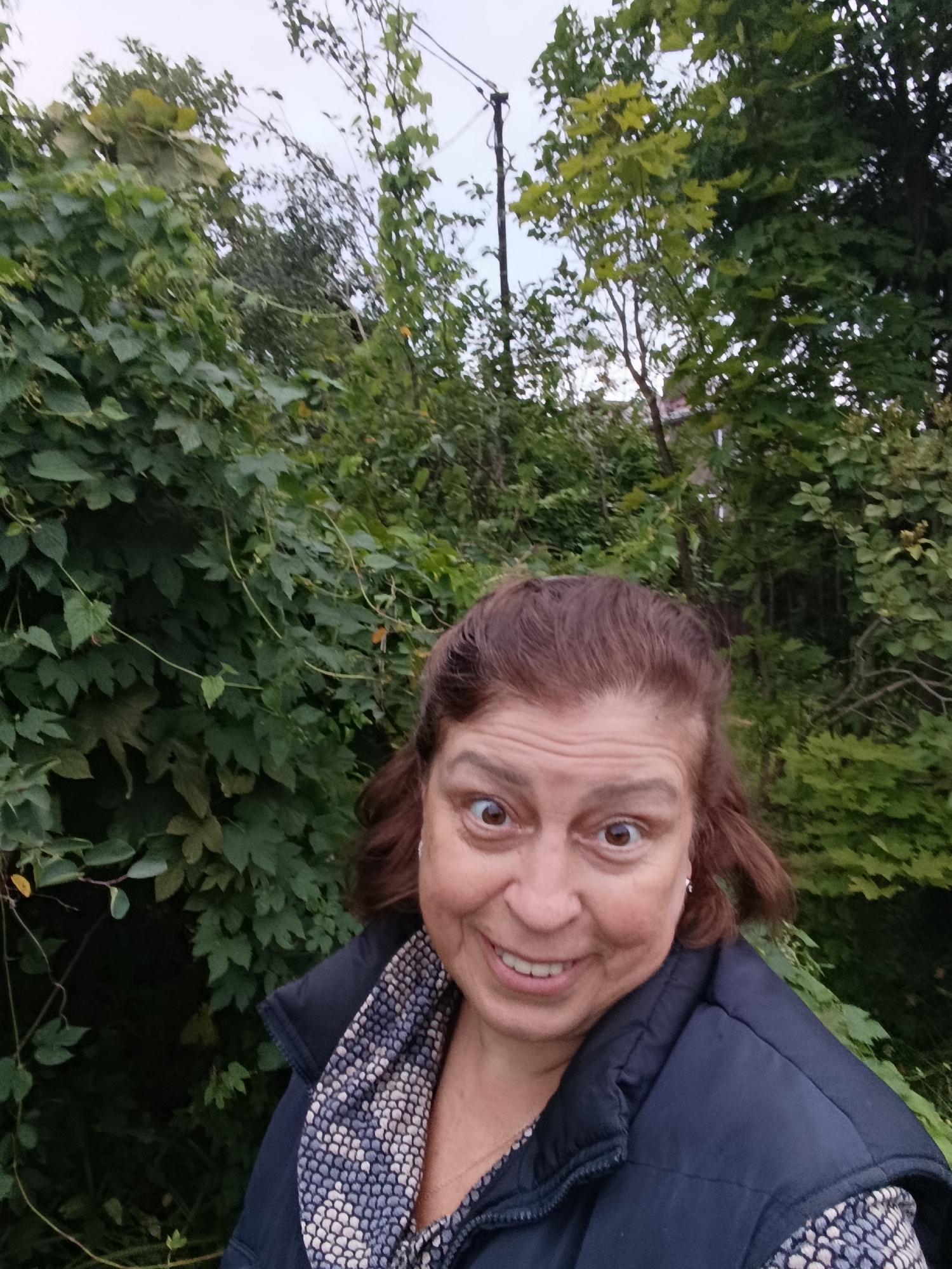 In the city of Verеya, Russia, a woman with brown hair and wide eyes smiles at the camera in a selfie. She is wearing a dark blue vest over a patterned shirt and a necklace. The background consists of lush green foliage and trees, with a hint of a utility pole and building in the distance. The lighting suggests it is late afternoon or early evening.