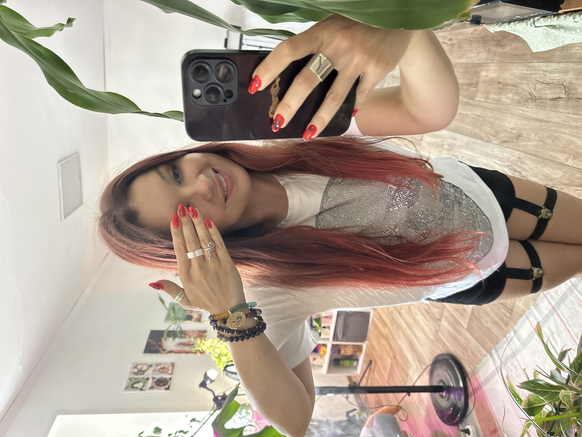A young woman with long, reddish-brown hair and red painted nails takes a selfie in a room with light-colored walls and a wooden floor. She is wearing a white t-shirt with a sparkly pattern, black shorts, and black garter straps around her thighs. She is covering one eye with her left hand, which is adorned with several rings. Her right hand holds a black smartphone, and her fingernails are painted red. In the background, shelves with various items, a fan, and a floor lamp are visible. A large green plant is to the right of the frame, partially obscuring the reflection of the room in a mirror. The image is consistent with an indoor setting in an urban environment, potentially a residential apartment in Vladimir, Russia, given the context.