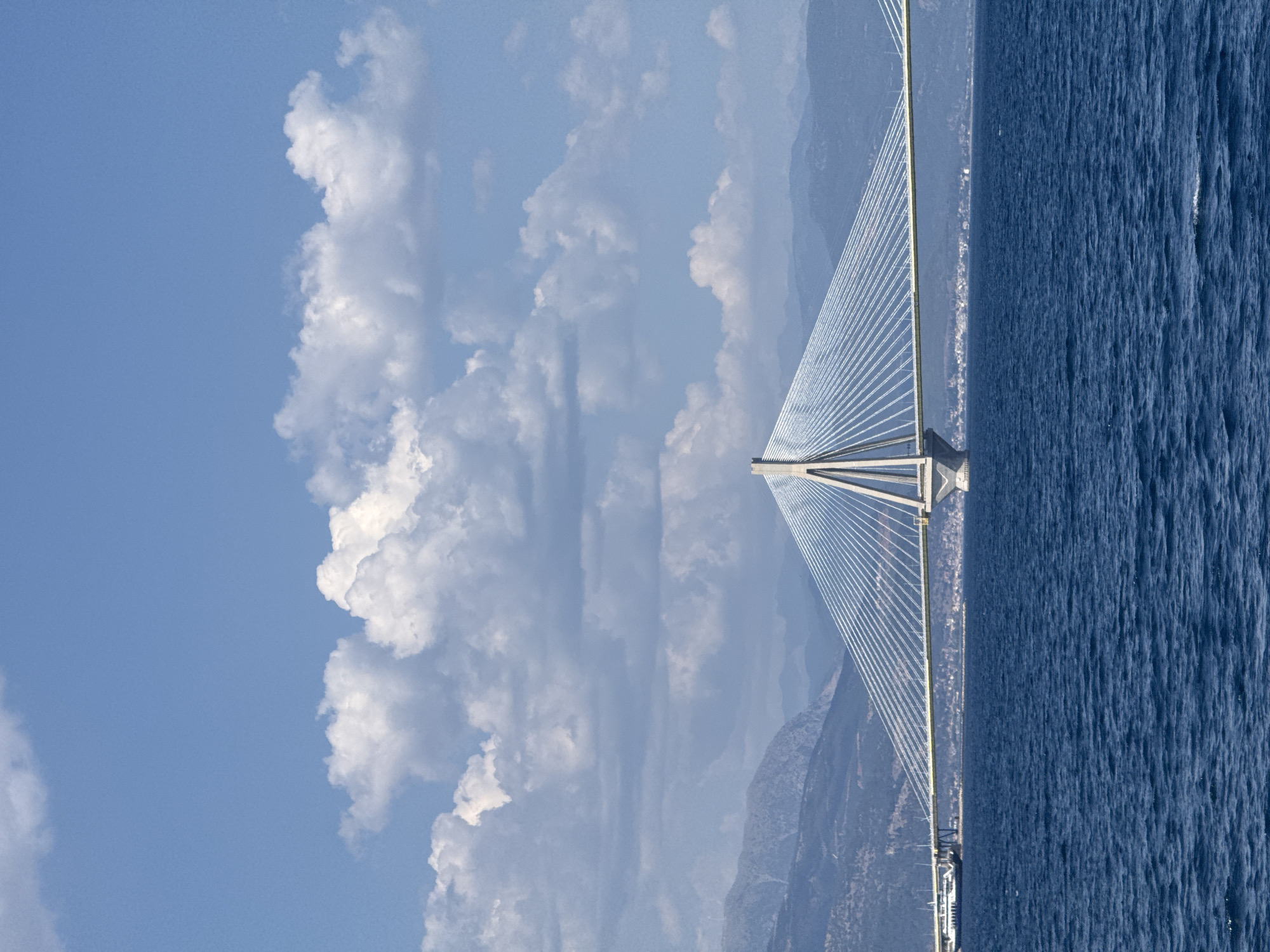This is a photograph of the Rio–Antirrio Bridge in Greece. The bridge, a prominent suspension bridge, stretches across a body of water that appears to be the Gulf of Corinth. The bridge's elegant design, with its numerous cables fanning out from a central tower, is a central subject. The water is a deep blue, with a textured surface suggesting gentle waves or ripples.
The background features a coastline with mountains. The mountains are muted in color, indicating distance and possibly a hazy atmosphere. The sky above is a bright blue, filled with fluffy white clouds, suggesting fair weather. The lighting indicates it is daytime, likely midday or early afternoon, given the brightness and the shadows.
The scene is serene and vast, with the bridge acting as a man-made structure connecting the natural elements of sea, sky, and land. There are no people visible in the frame. The overall impression is one of architectural achievement within a beautiful natural landscape. The photograph is oriented vertically, with the bridge running from the bottom left to the top right, and the sea occupying the right half of the frame.
