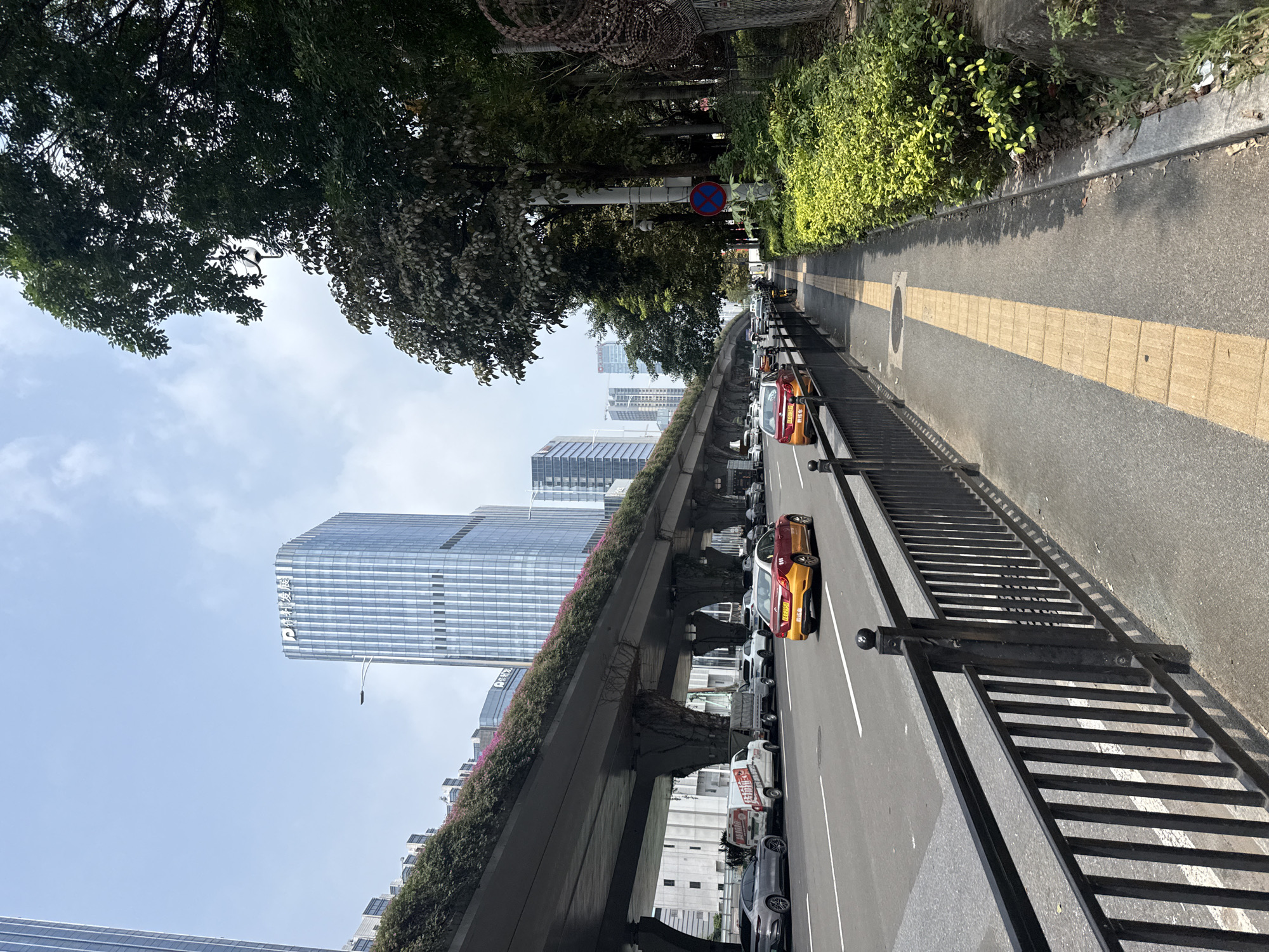 The image depicts a street scene in Huangpu District, China. A multi-level overpass, adorned with greenery and flowers along its edge, dominates the left side of the frame. Below the overpass, traffic moves along a wide asphalt road. Several cars are visible, including a red and yellow taxi, a silver sedan, a white van, and a small utility truck. On the right side of the road, a pedestrian sidewalk is bordered by a black metal fence. A yellow tactile paving strip runs along the sidewalk, indicating it is likely a pathway for visually impaired individuals. Lush green bushes and trees line the sidewalk further to the right, with a concrete curb separating the vegetation from the walkway. In the background, modern high-rise buildings with glass facades rise into a clear blue sky with scattered clouds. A street lamp and a security camera are also present on a pole along the sidewalk. The overall scene suggests a bustling urban environment with a combination of elevated infrastructure and street-level activity.