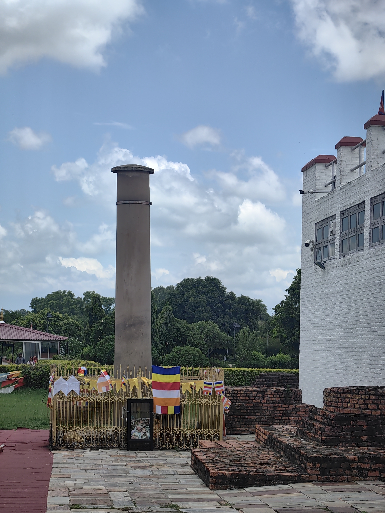 In Lumbini Bazaar, Nepal, a tall, gray, cylindrical pillar stands prominently against a cloudy blue sky. To its right, a white brick building with red-capped architectural features and dark-paned windows is visible, along with several tiered brick structures in the foreground. A golden, ornate fence surrounds a display case containing relics, adorned with prayer flags in various colors, including a large Tibetan flag. In the background, lush green trees and a red-roofed pavilion with people can be seen. The ground is paved with stone tiles, with a red path leading towards the left side of the frame.