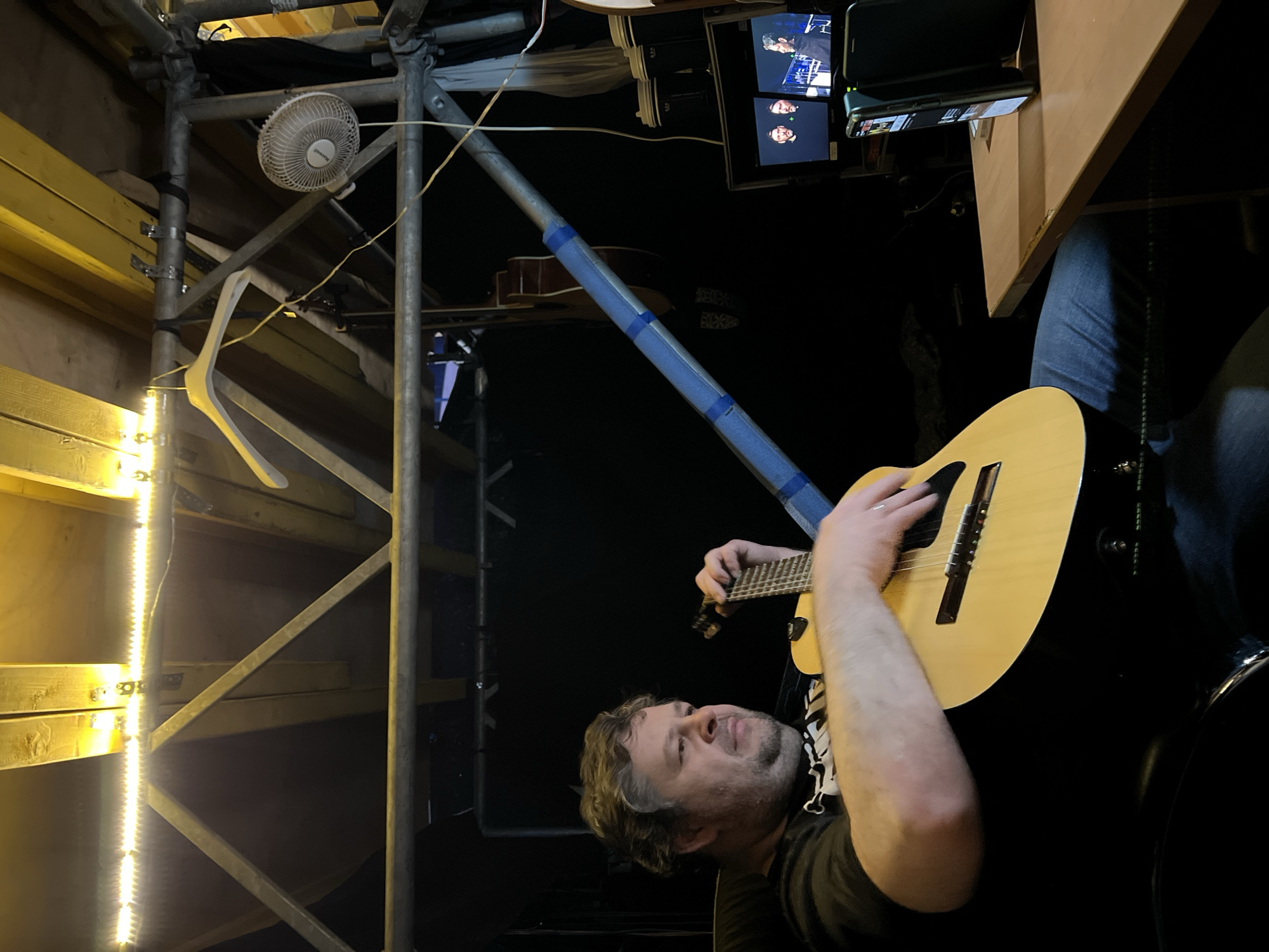 The image shows a man playing a guitar in what appears to be a makeshift or backstage area, possibly in Khimki, Russia. The man is in the lower right foreground, and he is in focus. Behind him, there is scaffolding with a string of lights attached. The scaffolding also has a small fan and a clothes hanger. Above the scaffolding, there is a monitor with multiple screens showing what appears to be video footage of people, and a desk holding a smartphone and some electronics. A blue-taped pipe runs diagonally across the middle of the frame. The overall lighting in the area is dim and artificial.