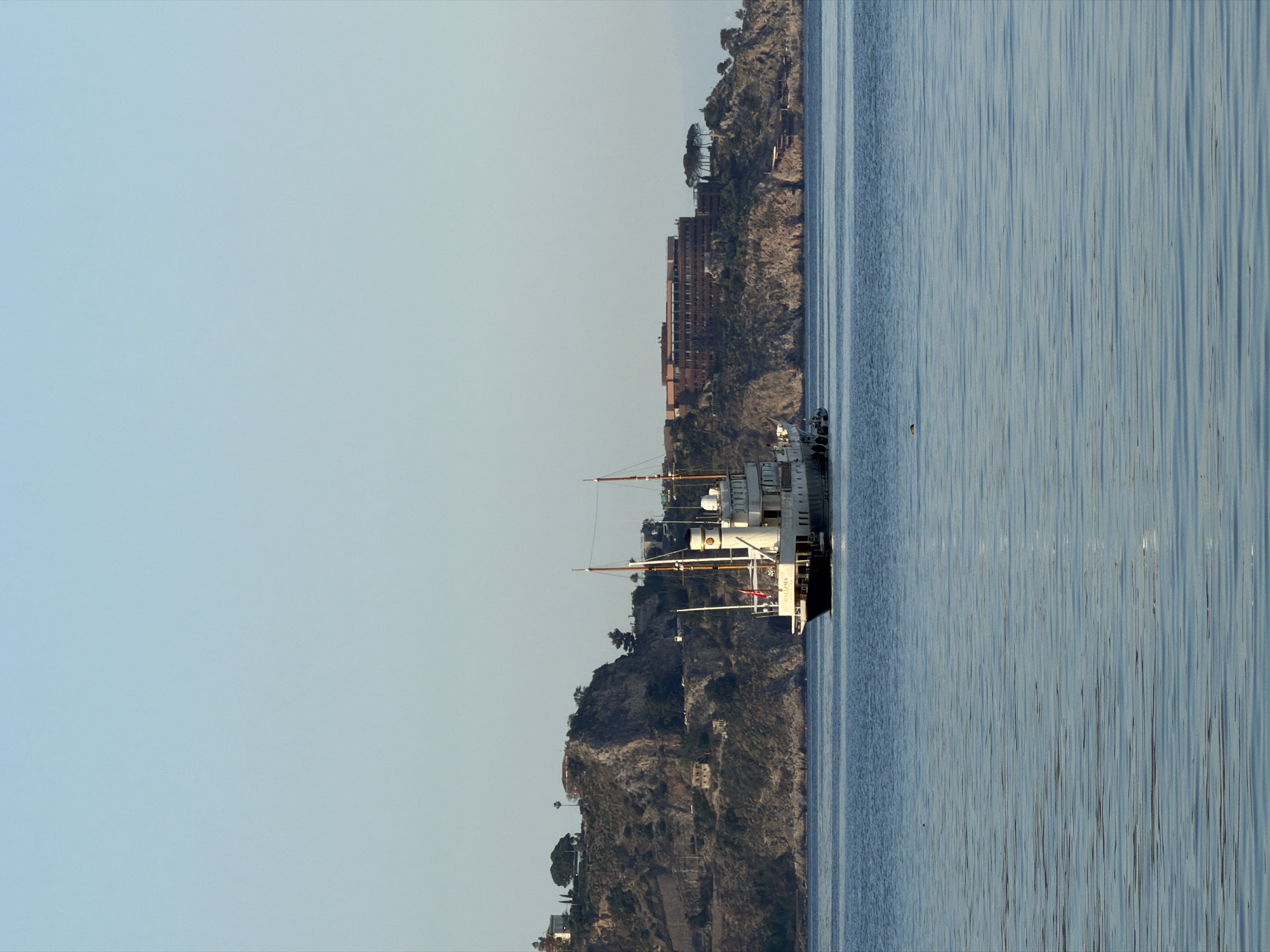 The image shows a sideways view of a body of water, likely the Ionian Sea near Chianchitta, Italy, with a ship docked along the coast. The shoreline consists of rocky cliffs with vegetation and buildings. A large, multi-story hotel is also visible. The sky is clear and light blue. The water is calm with subtle reflections.