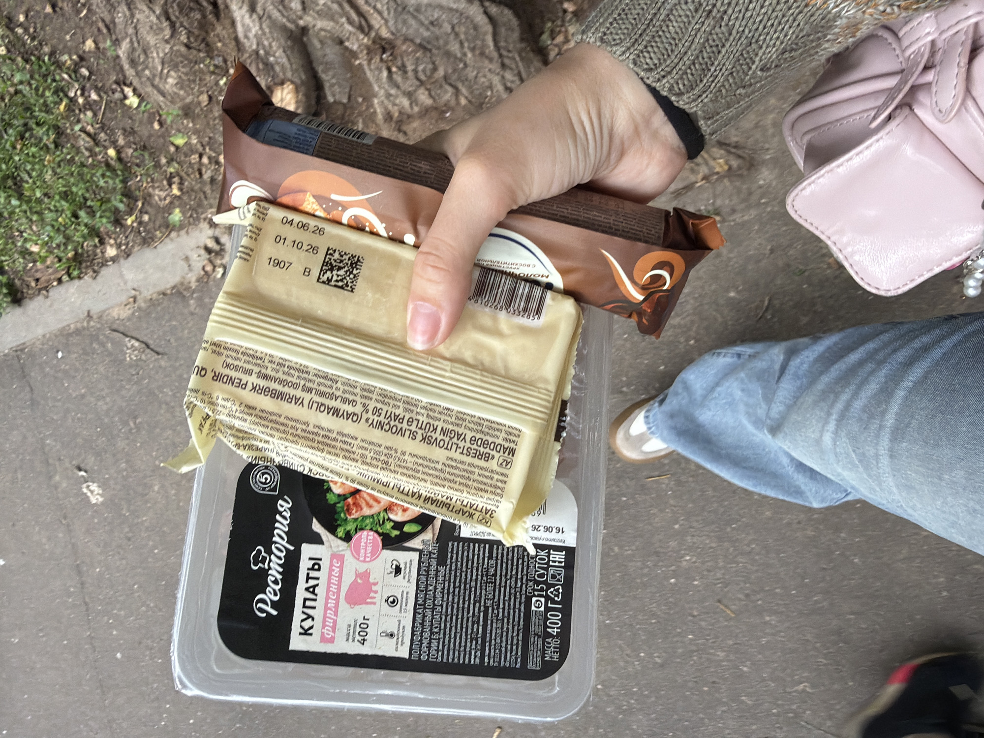 In a close-up shot, a person's hand holds a selection of food items. The dominant items are two chocolate bars and a package of "Рестория" brand meat products, specifically "КУПАТЫ фирменные" (Kupati, branded). The meat package is a clear plastic container with a black label featuring an image of cooked meat, the brand logo, and text in Russian. The expiration dates visible on the chocolate bars are 04.06.26 and 01.10.26. The meat product's expiration date is 16.06.26, and it is marked with a 5-day expiration after opening. The setting appears to be outdoors, with the ground showing a mix of asphalt and dirt with some sparse vegetation and tree roots visible. The person is wearing a grey knit sweater and blue jeans, and a light pink purse is partially visible. The lighting suggests it is daytime. The scene captures a moment of someone holding groceries.