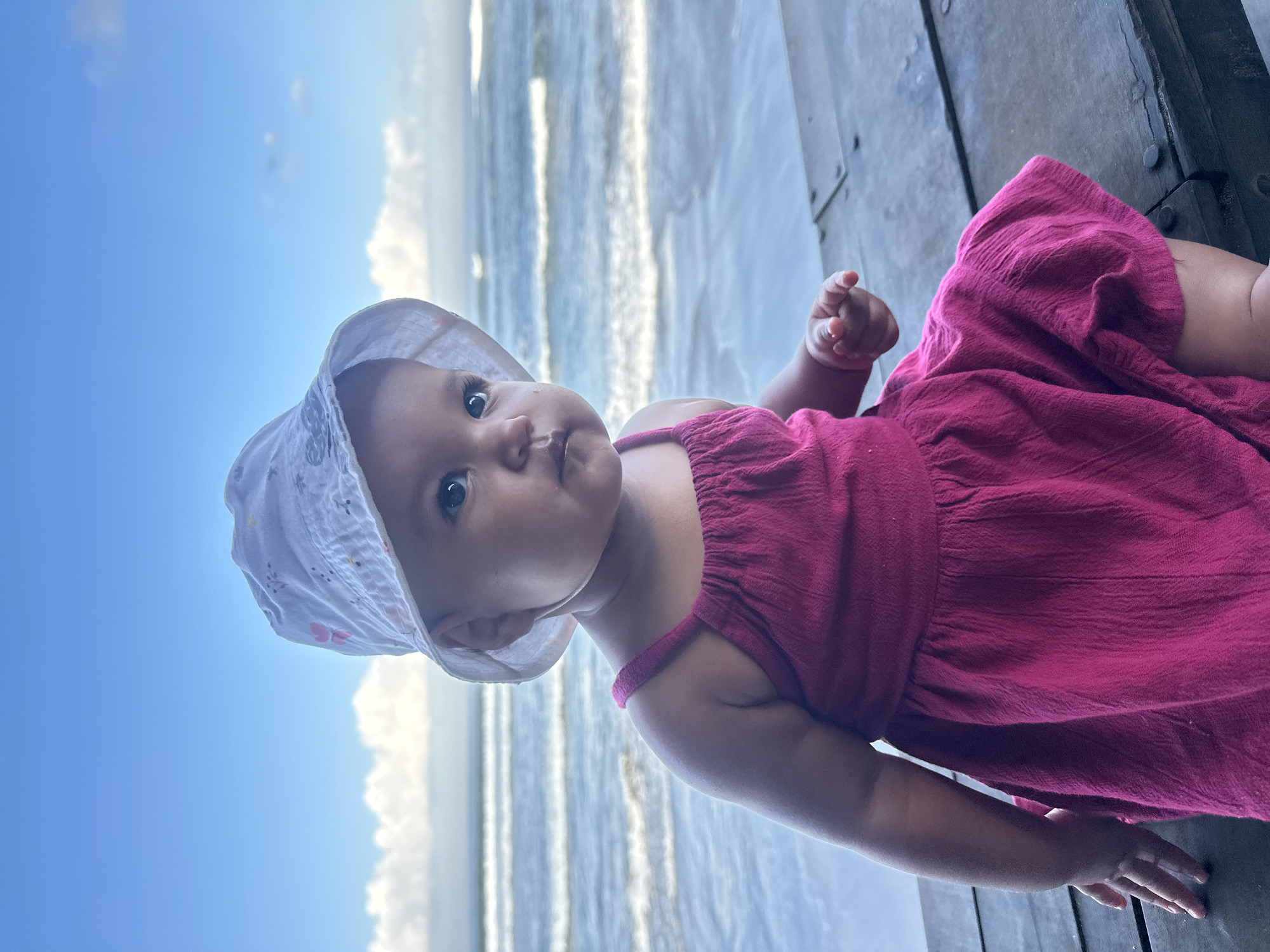 An outdoor, eye-level, medium shot shows a baby girl sitting on a wooden surface with the ocean in the background. The baby is wearing a white bucket hat with pink flowers and a pink sleeveless dress. She is looking to her right with her mouth slightly open. The ocean has several waves with white crests. The sky is blue with some white clouds. The wooden surface appears to be a pier or deck. The location is North Kuta, Indonesia.