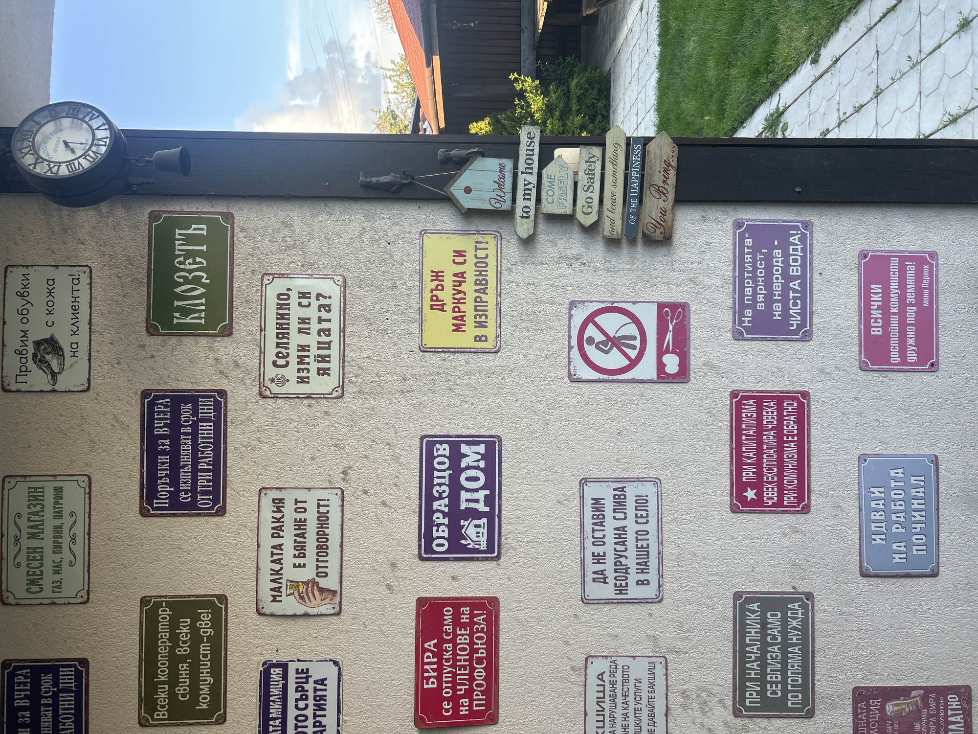 The image depicts a wall covered in signs written in Bulgarian, situated outdoors. A decorative clock is mounted above the signs. The signs display various humorous or satirical messages about everyday life, work, politics, and social norms, typical of a pub or restaurant interior. Several signs also feature small graphics or illustrations related to their text. A series of small wooden signposts with messages like "Welcome to my house," "Go Safely," and "You Bring..." are affixed to the top of the wall. The top of the wall is covered by a black frame. The image suggests the location could be a public house in the village of Konska, Bulgaria, based on the language and cultural references in the signs. The environment seems to be an outdoor seating area or a courtyard.