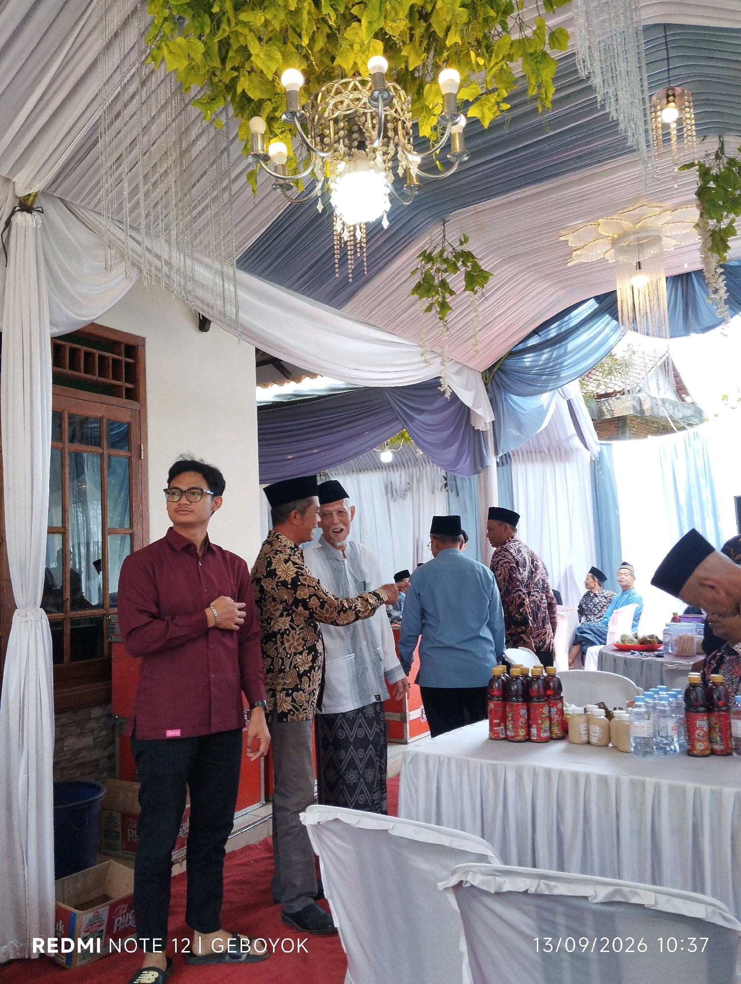 The image depicts an outdoor event, possibly a wedding or a similar celebration, taking place under a decorated tent in Kuwukan, Indonesia. The tent's ceiling is adorned with draped white and blue fabric, green foliage, and chandeliers. In the foreground, a young man wearing glasses and a maroon long-sleeved shirt and black pants stands looking to his left. To his right, two older men in traditional attire, one in a patterned batik shirt and the other in a white shirt and checkered sarong, are engaged in conversation, with the older man on the right gesturing. Further into the background, several other individuals are seated and standing, many wearing peci caps, indicative of a Muslim gathering. A table laden with bottled drinks and other items is visible, draped with a white tablecloth. The overall atmosphere suggests a community event in an urban or semi-urban setting in Indonesia.