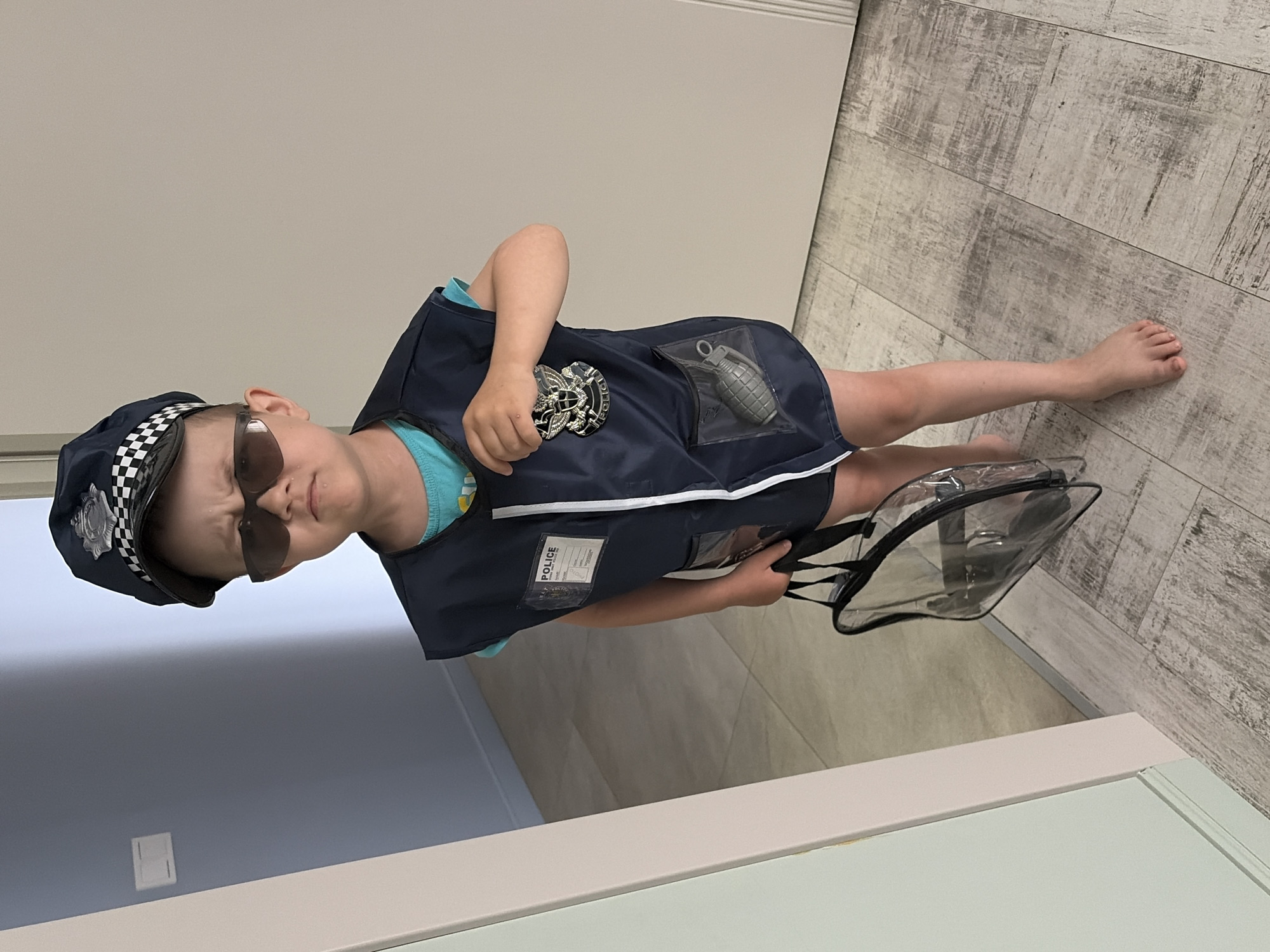 A young child dressed in a police officer costume stands indoors. The child wears a police cap, sunglasses, and a dark blue vest adorned with a police badge and a simulated grenade in a clear pocket. The child holds a clear bag with black straps. The background includes a wall painted in light shades of gray and blue, and a light-colored wooden floor. The image does not contain any information to indicate the location of Chaikovsky, Russia.
