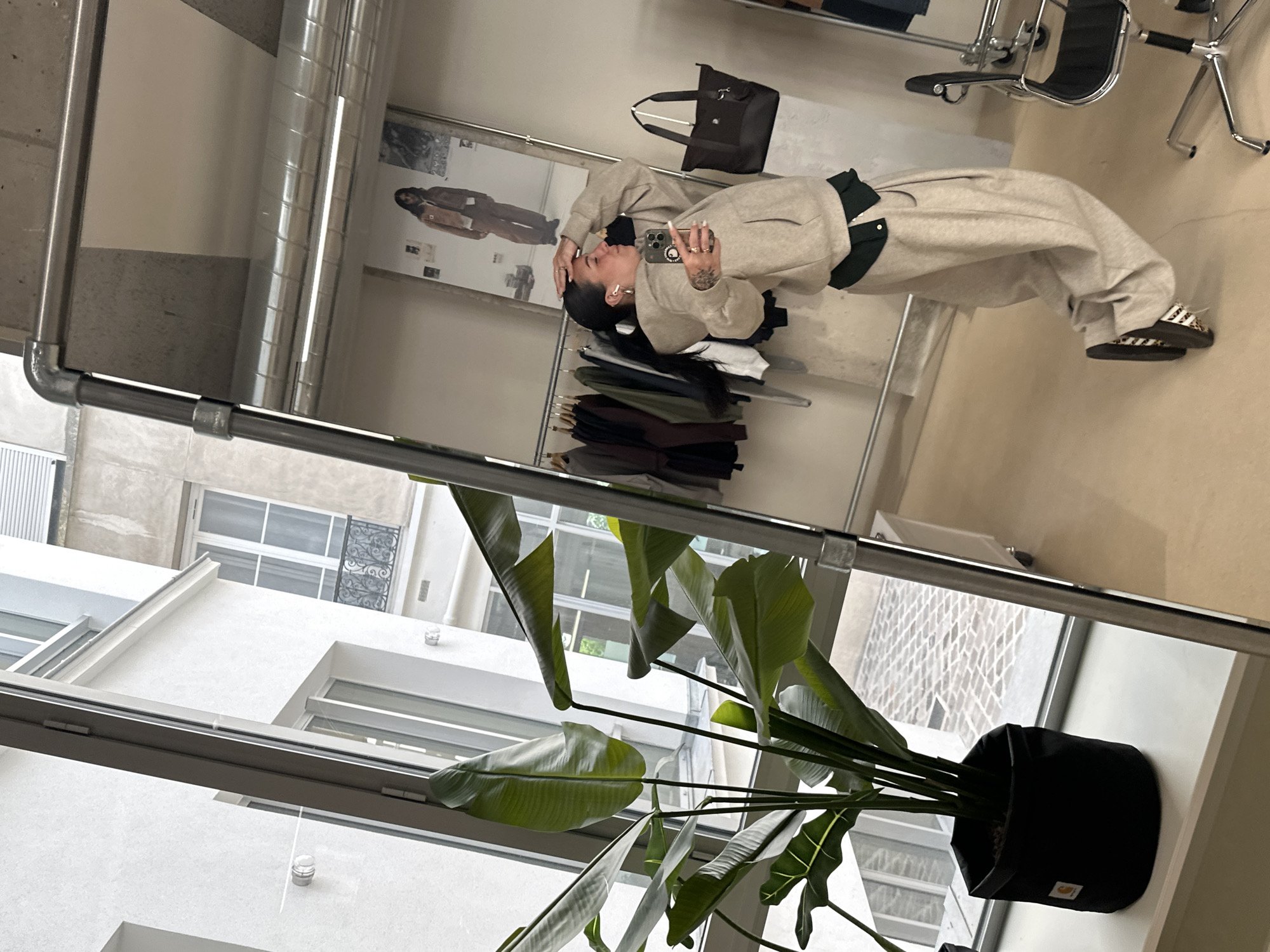 The image shows a reflection of a room interior in a mirror. A woman lies on a surface, dressed in a beige ensemble. Next to her is a rack of clothing, and a bag is placed nearby. Seated chairs are also visible in the background. The mirror reflection also reveals an exterior view of a building with multiple windows. In the foreground, there's a houseplant in a black pot placed on a windowsill. The architectural details visible through the windows are typical of buildings in Paris, France. The presence of modern architectural elements suggests that the location could be a contemporary commercial or residential space in Paris.