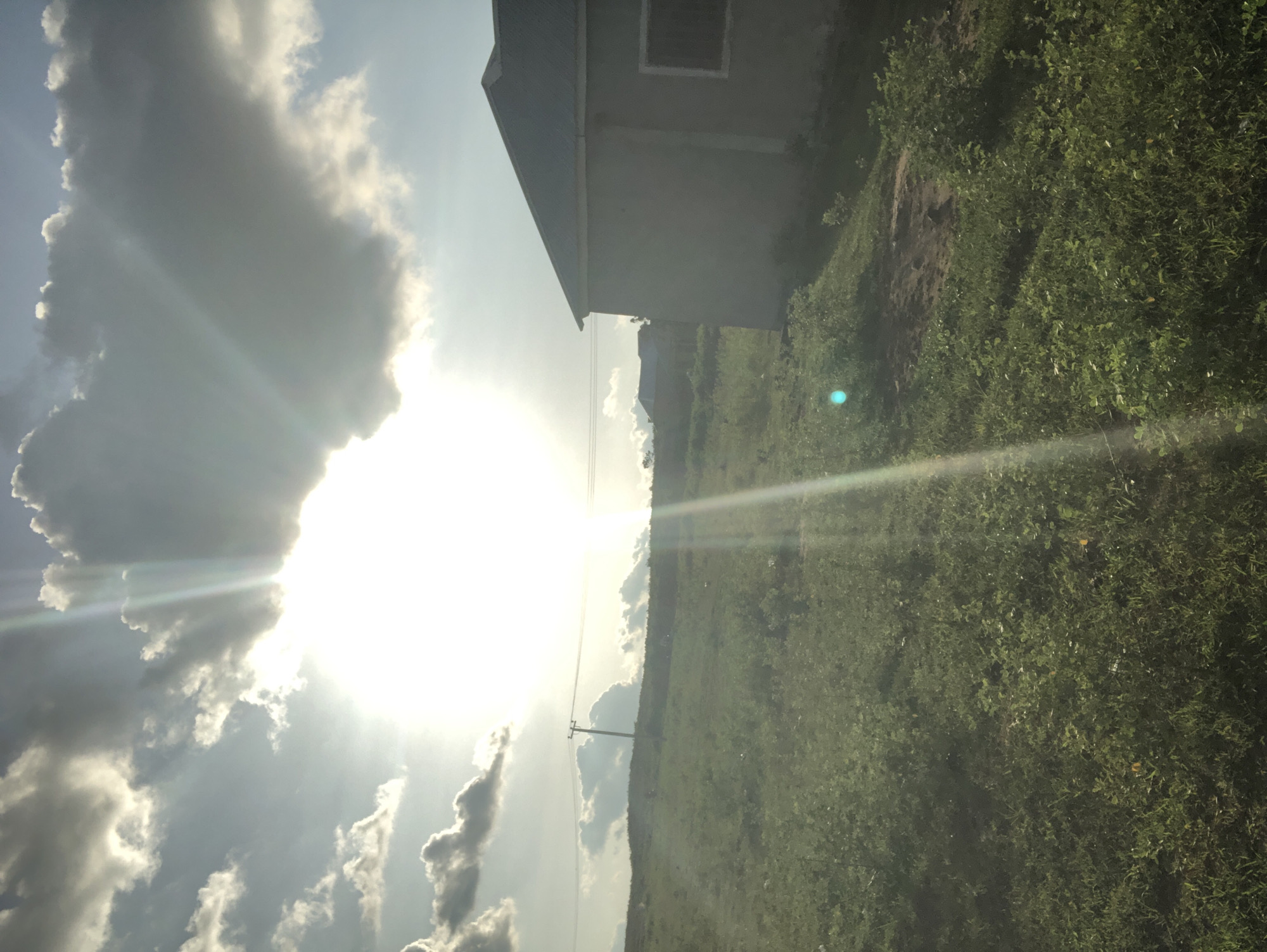 In an outdoor scene in an unspecified location in Nigeria, the sun shines brightly through scattered clouds, casting rays of light across the landscape. A portion of a light-colored building with a dark roof is visible on the right side of the image. The ground is covered with green vegetation, including grass and bushes. In the distance, a utility pole with wires stands on a grassy hill. Another building with a red roof is also visible on the hill. The sky is partly cloudy with dramatic light effects from the sun.