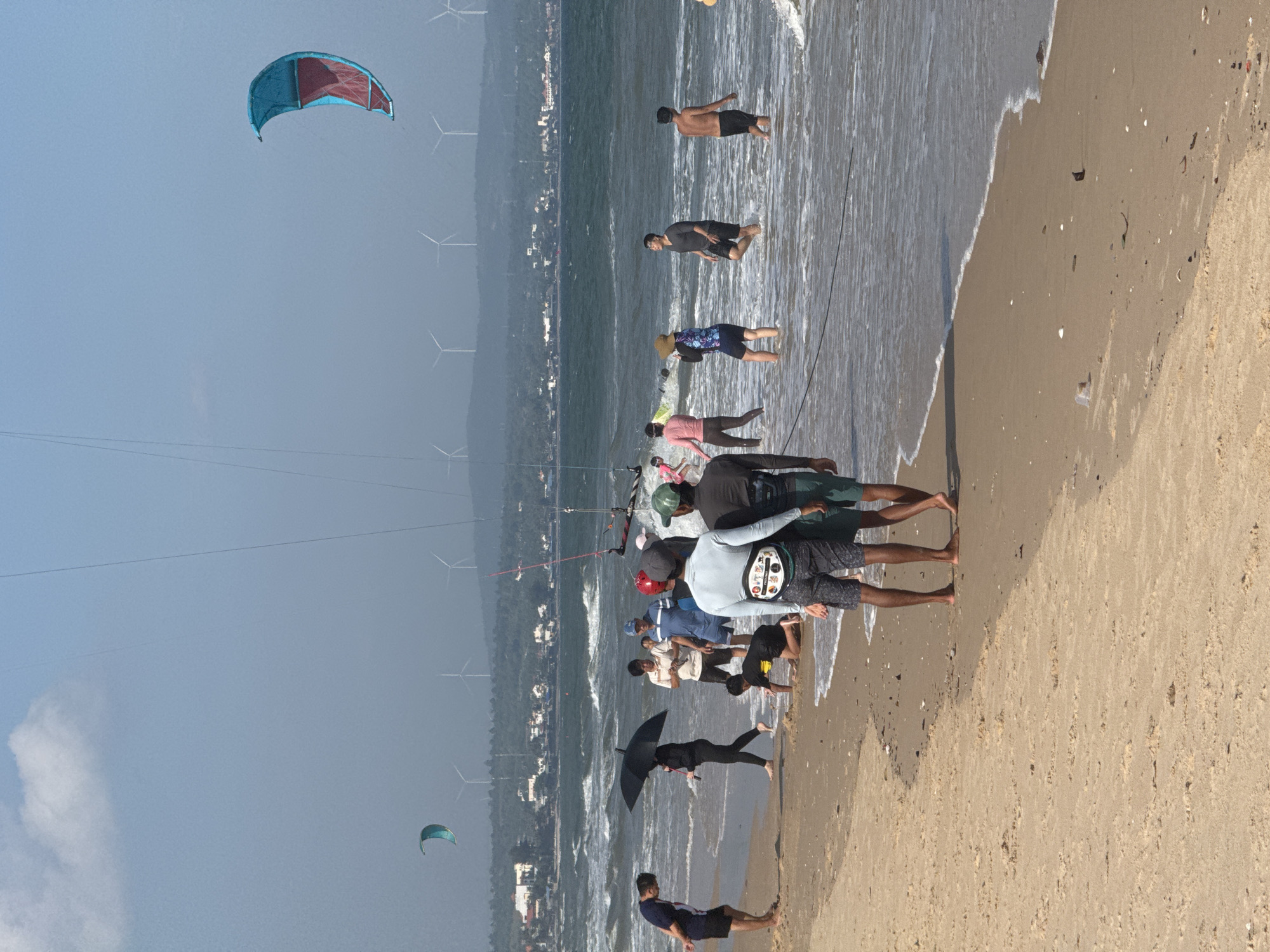 The image depicts a bustling beach scene in Mũi Né, Vietnam, likely during daylight hours given the bright sky. A group of people are engaged in or observing kiteboarding activities. In the foreground, several individuals stand on the sandy shore, some wading in the shallow water. One person in the center is holding a black umbrella. Further back, two men are walking in the water. In the distance, a large, colorful kite is visible in the sky, along with smaller kites closer to the water. Wind turbines dot the horizon, suggesting a location with consistent wind. The ocean is wavy, indicating moderate to strong winds. The background shows a coastline with buildings and a hazy landscape, consistent with the coastal environment of Mũi Né.