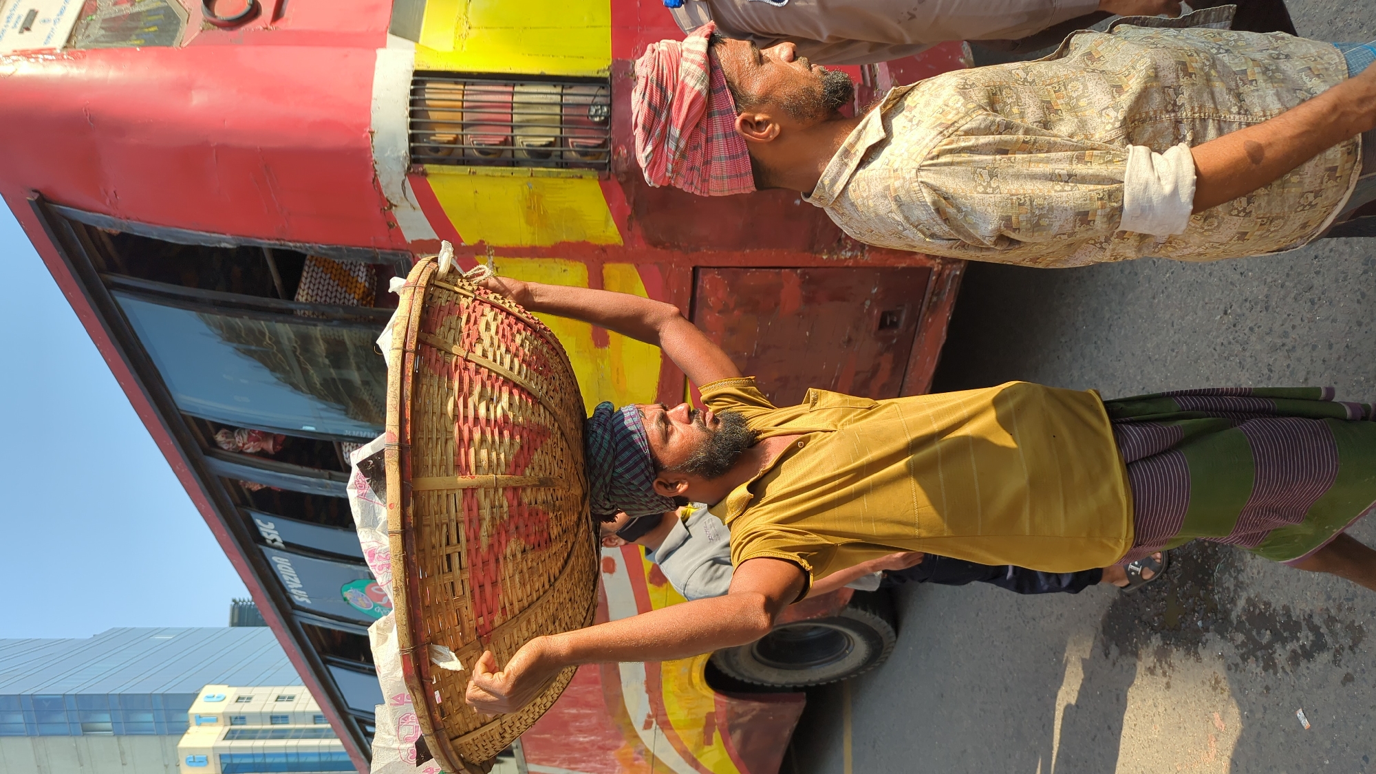 In Dhaka, Bangladesh, a man carries a large, woven basket on his head while standing next to a red and yellow bus. He wears a yellow shirt, a striped cloth around his waist, and a checkered head covering. Another man stands nearby, wearing a patterned shirt and a red and white checkered head covering. The background includes a modern building and glimpses of people inside the bus. The scene is set on a street during the day.